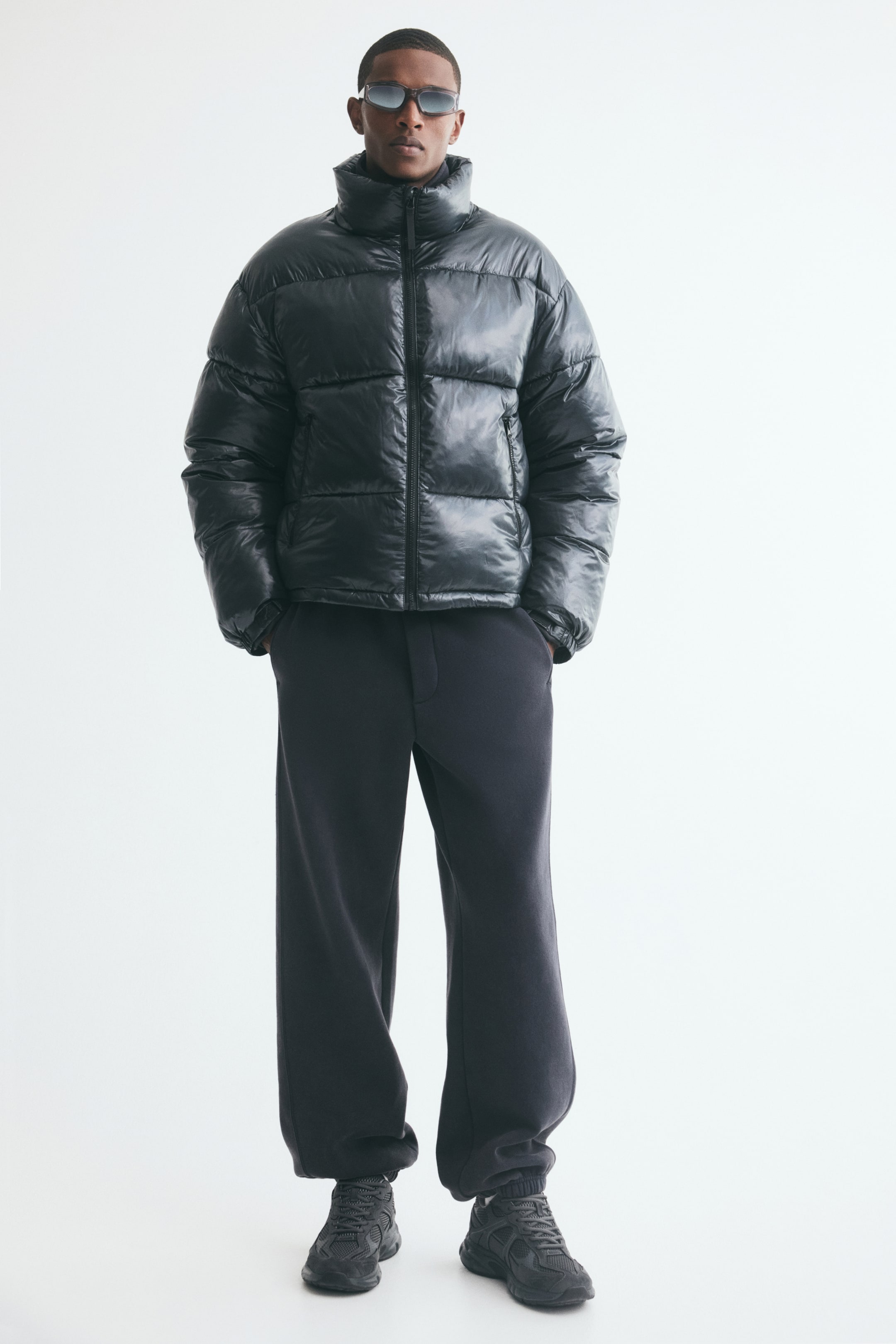 Voir une image plus grande: Un blouson matelassé noir à l’aspect lustré, doté d’un col montant et d’une fermeture éclair à l’avant, est porté par le mannequin. Le blouson est stylé avec un pantalon foncé ample et des espadrilles assorties.