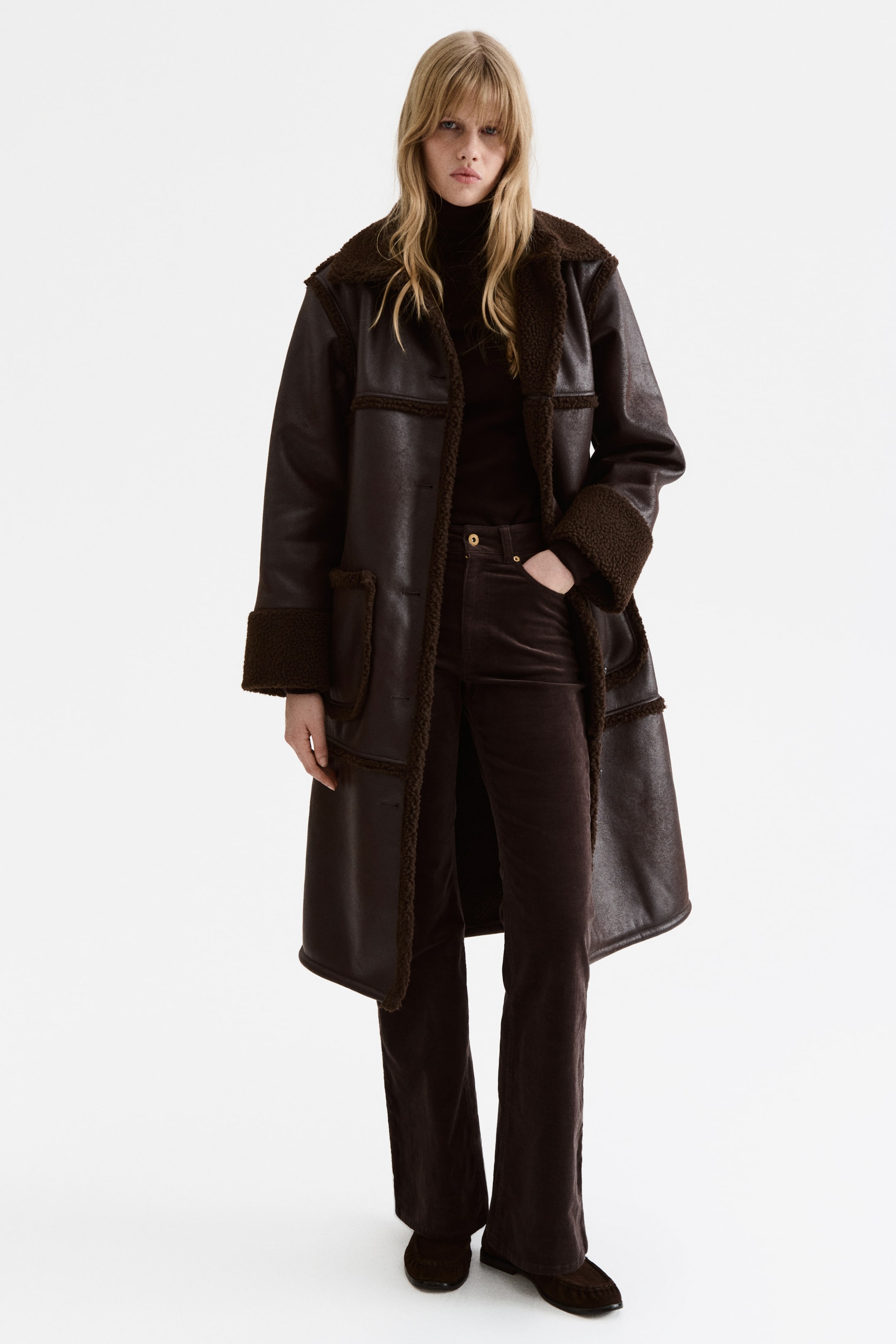 Größeres Bild anzeigen: 5-Pocket-Hosen aus weichem Cord werden vom Model getragen. Diese dunkelbraunen Hosen haben eine hohe Taille mit sichtbarem Reißverschluss und Knopf, eine schlanke Passform und ausgestellte Beine. Die rechte Hand des Models ruht in einer Vordertasche.