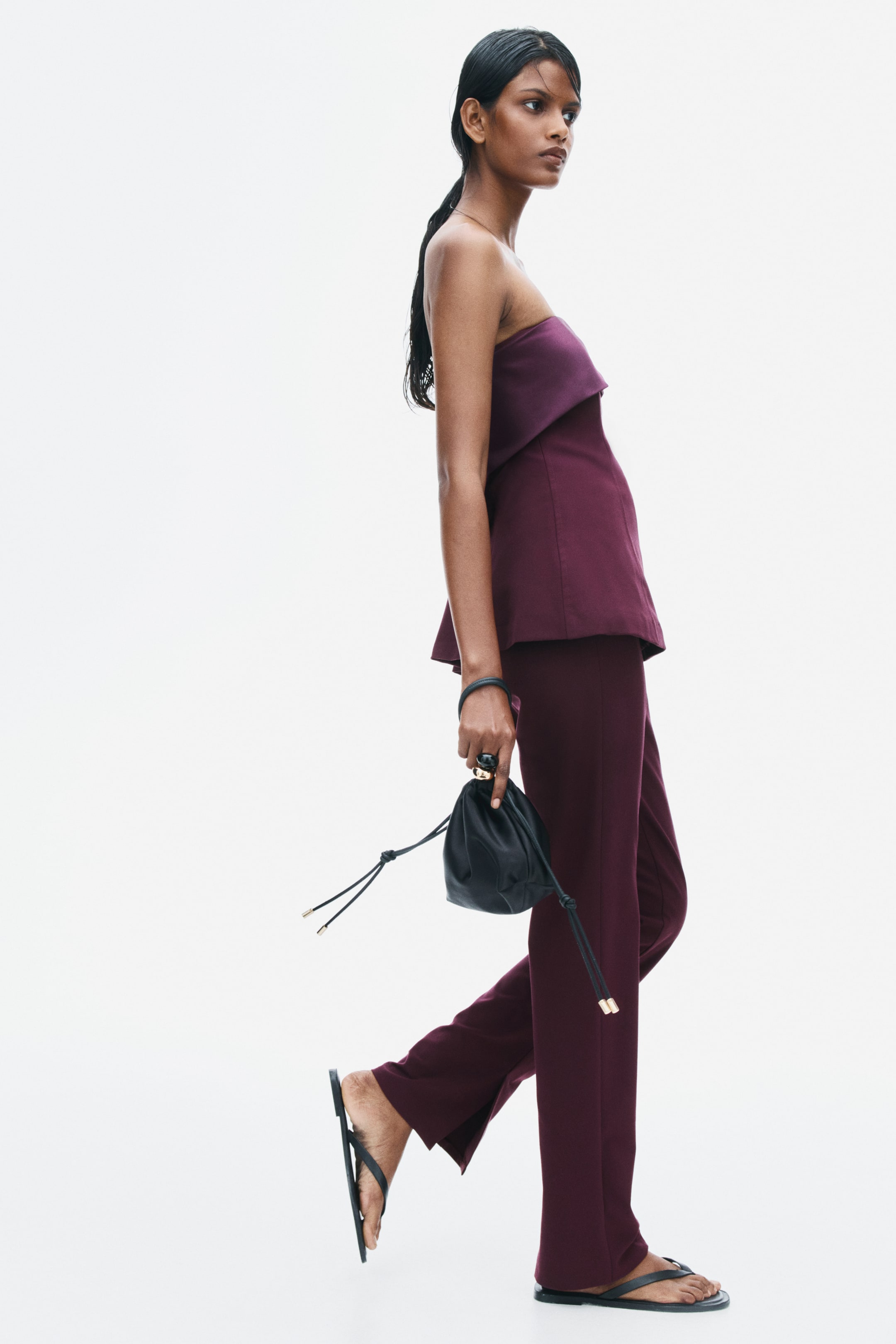 View larger image: Burgundy fitted pants in a woven fabric are being worn by the model, featuring a slim-leg silhouette with a slit at the side hem. The pants are styled with a matching burgundy strapless top, flat black sandals, and a small black drawstring handbag.