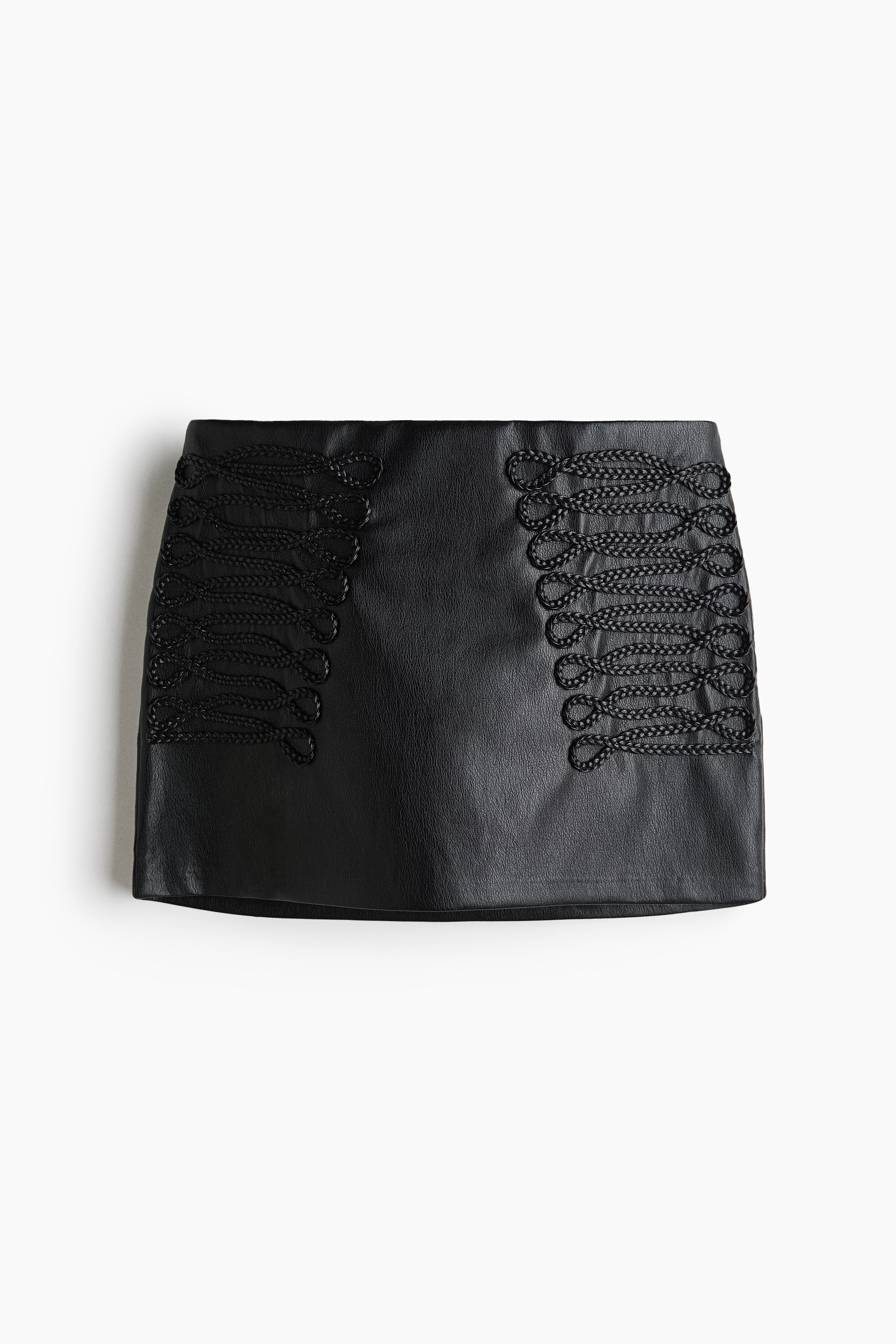La modelo lleva una falda corta negra de tejido recubierto, con detalles decorativos de cordÃ³n trenzado a ambos lados del delantero. La falda se combina con una blusa blanca de manga larga de volumen y medias negras transparentes.