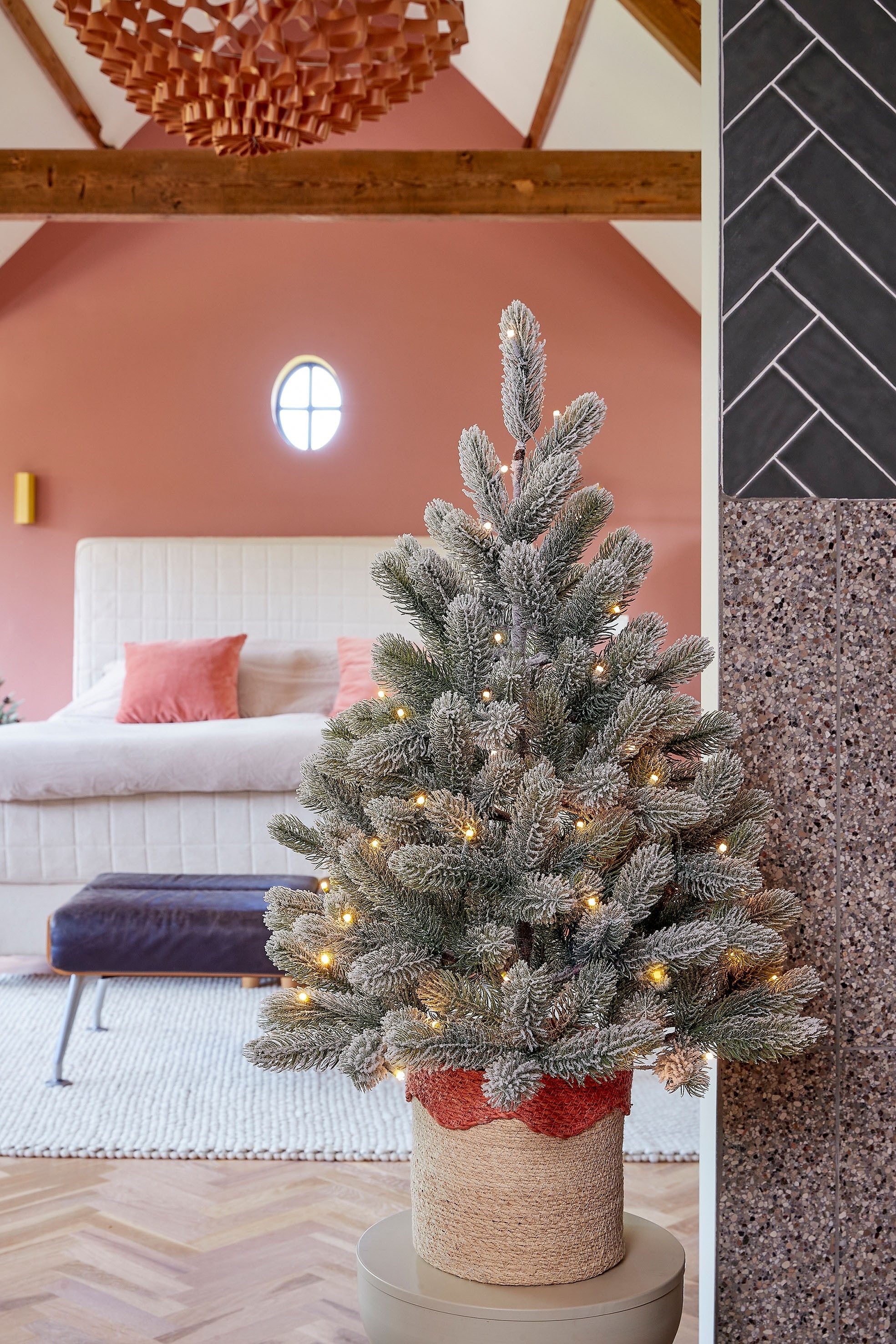 Ein künstlicher Weihnachtsbaum steht frontal, seine grünen Zweige sind mit weißem Flock besetzt und mit kleinen gelben Lichtern integriert, und er wird in einem schwarzen Topf präsentiert.