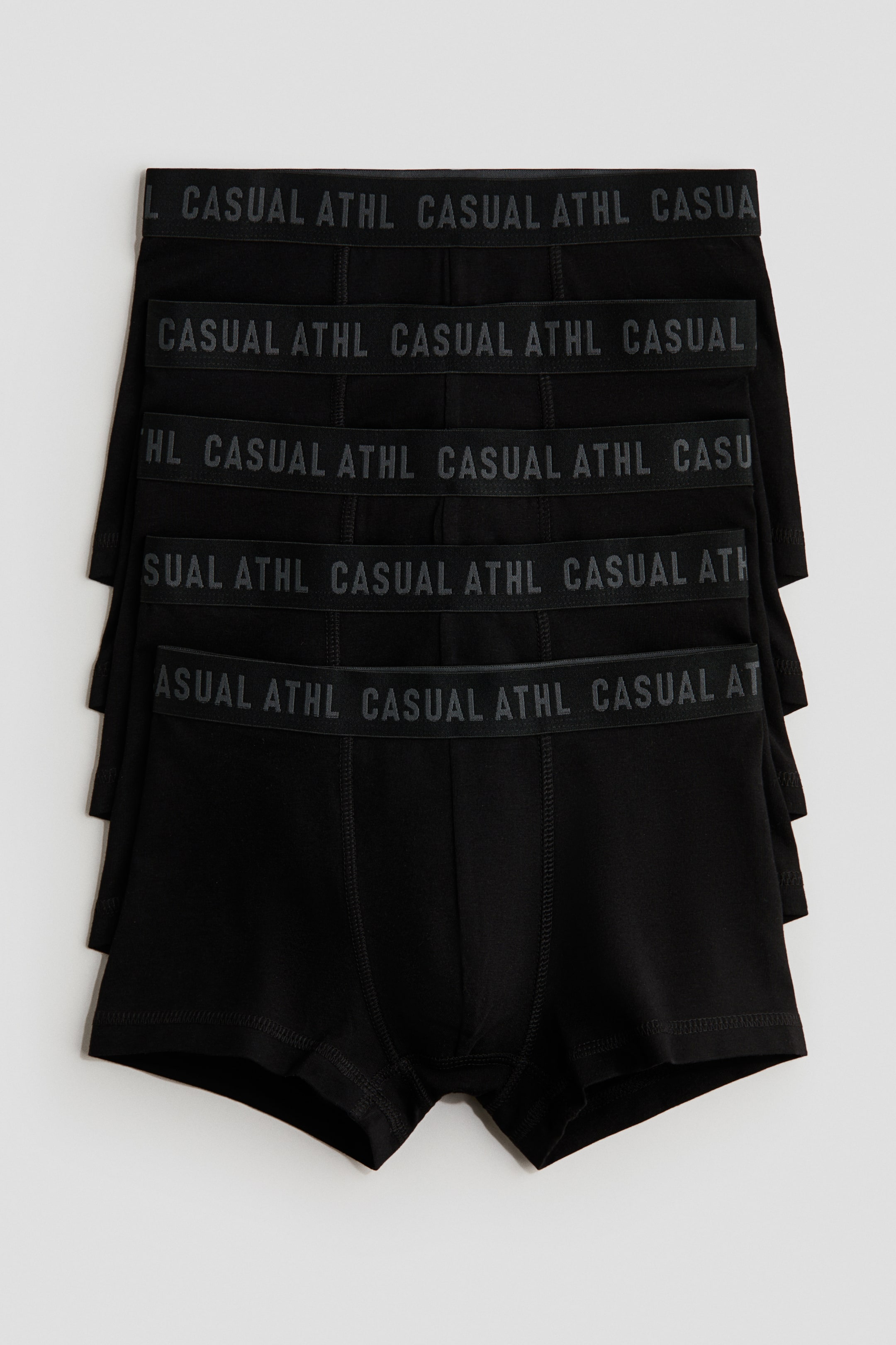 View larger image: Five black boxer briefs are stacked and fanned out, each pair featuring a black elastic waistband with repeating gray "CASUAL ATHL" text. The visible front of the briefs displays a plain black body with prominent front panel stitching.