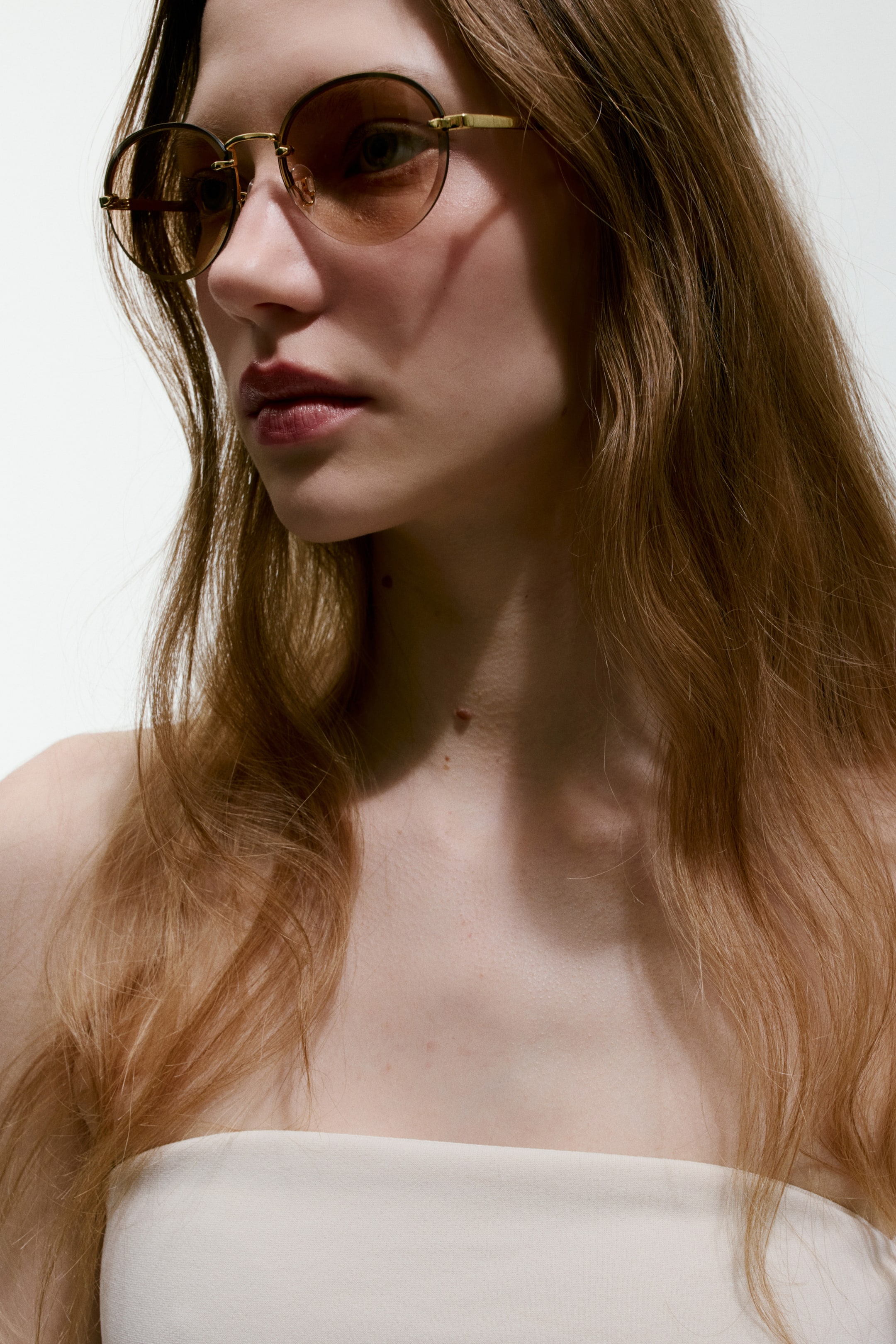 View larger image: The sunglasses feature round, brown-tinted lenses set in a slim, gold-toned metal frame. They are shown being worn, angled slightly to the right, highlighting the metal temples extending backward.