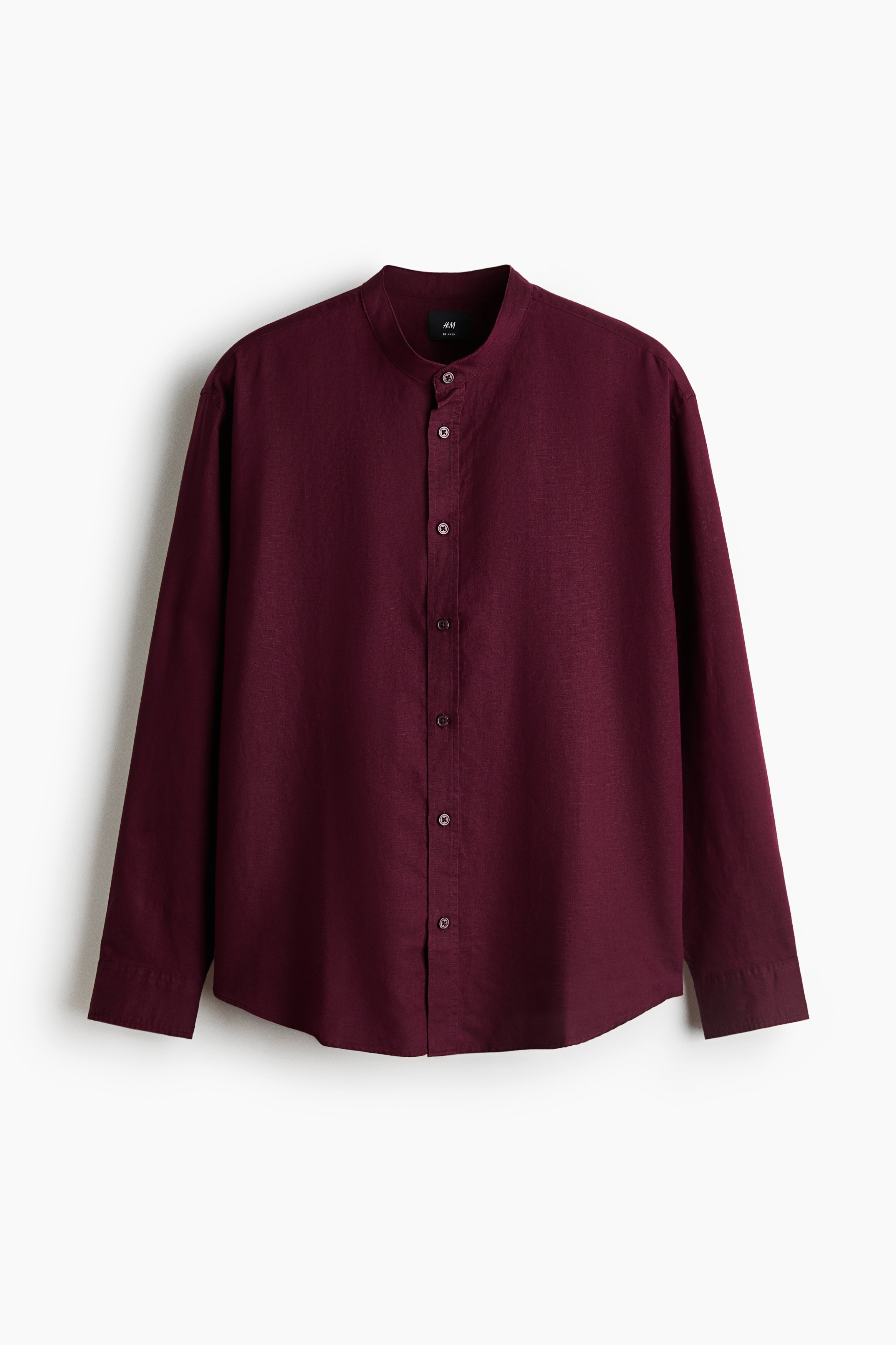 A burgundy linen-blend shirt featuring a band collar, button-up front, and long sleeves is being worn by the model. The relaxed-fit shirt is styled untucked over wide-leg black trousers, while the model is carrying a black textured tote bag.