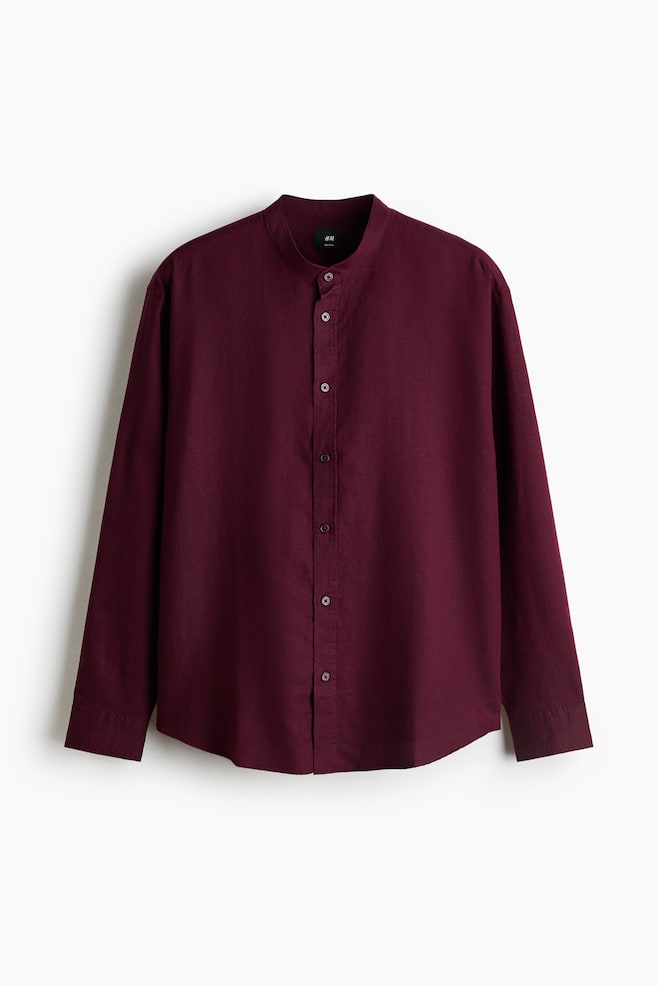 A burgundy long-sleeve shirt in a textured linen-cotton blend is displayed facing forward, featuring a band collar, a button-down front, and a rounded hem. The garment is designed with a relaxed fit and long sleeves ending in buttoned cuffs. - 1