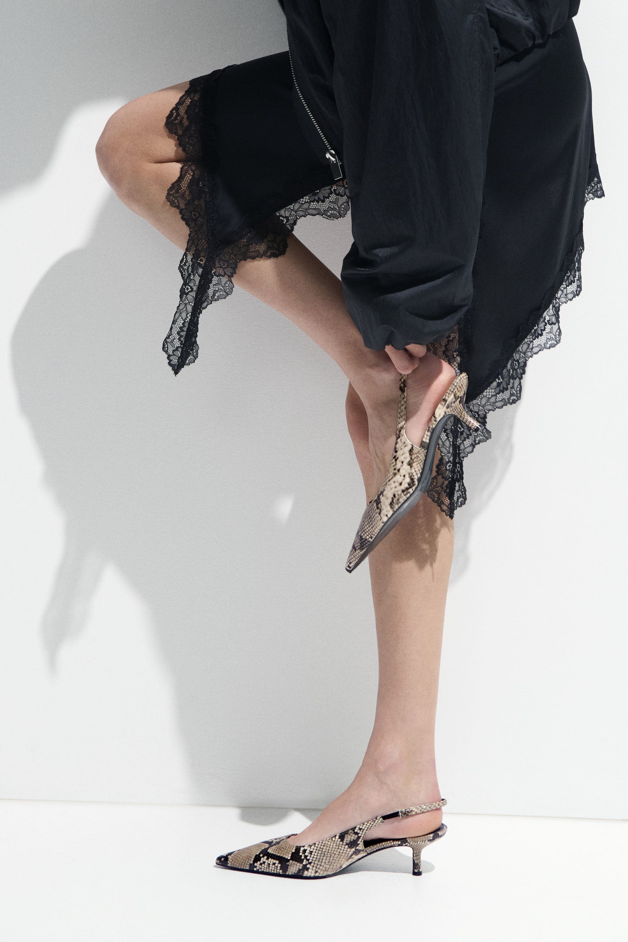 View larger image: A black slip garment featuring a satin-like texture, intricate black lace trim along its scalloped hemline, and a visible side zipper, draping fluidly over a raised leg.