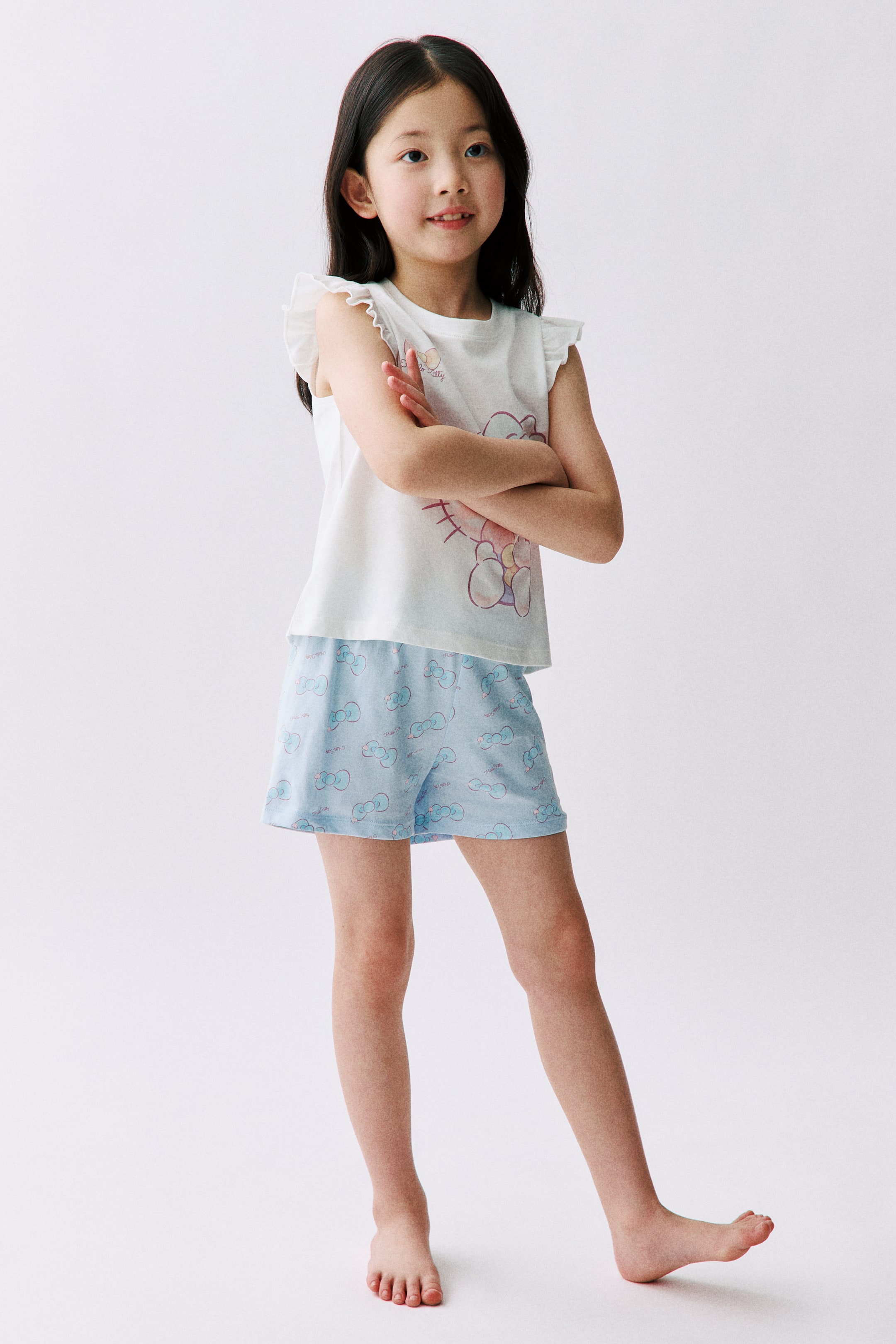 View larger image: Pyjamas with a white cotton jersey top showcasing a graphic print, a round neckline, and flutter sleeves, paired with light blue cotton jersey shorts featuring an all-over print and an elasticated waist. The model is wearing the pyjamas, standing with arms crossed.