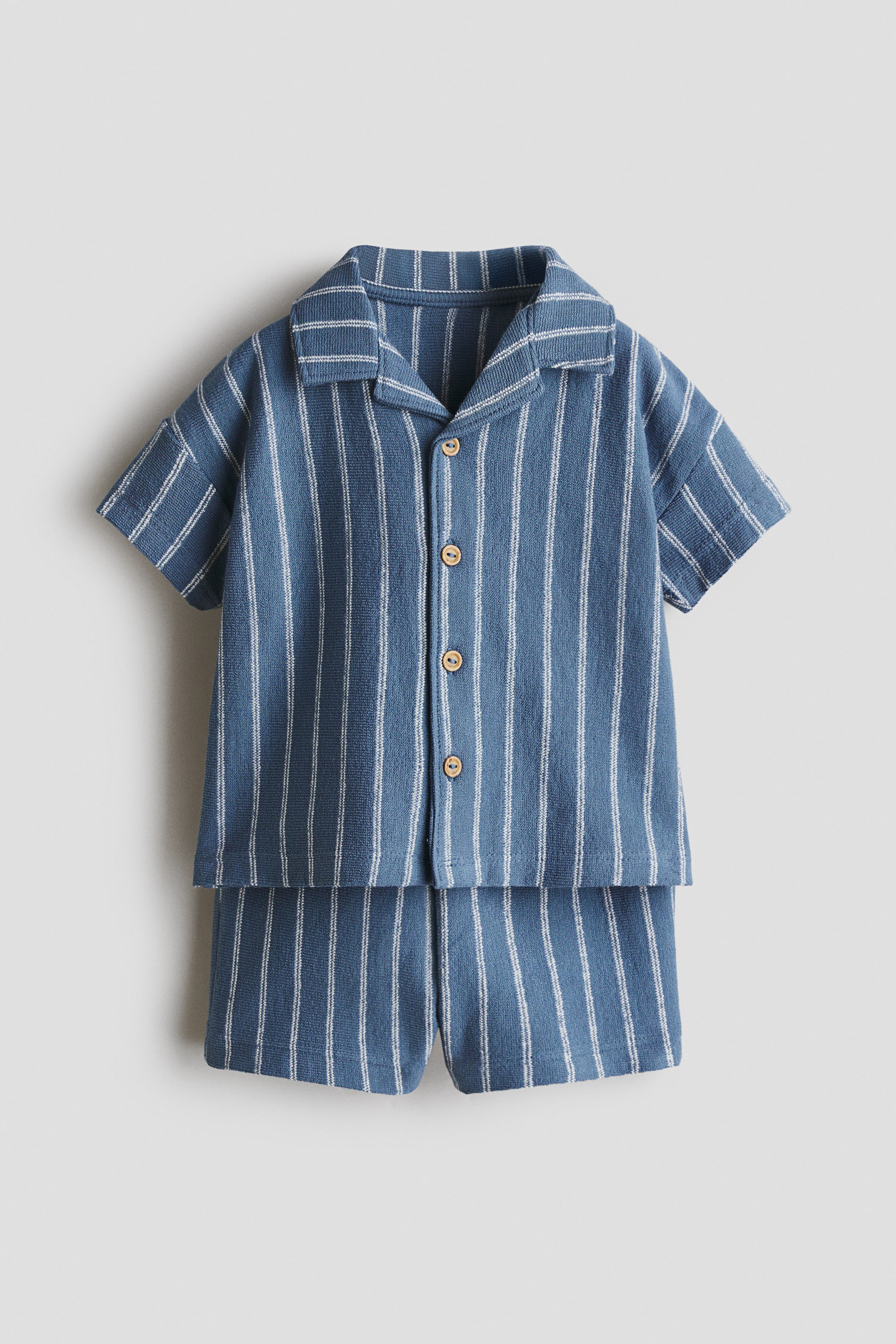 View larger image: A blue two-piece set, consisting of a short-sleeved top and shorts, is laid flat and shown from the front. The set is made of textured knit fabric with white vertical stripes, and the top features a spread collar and wood-look buttons down the front.