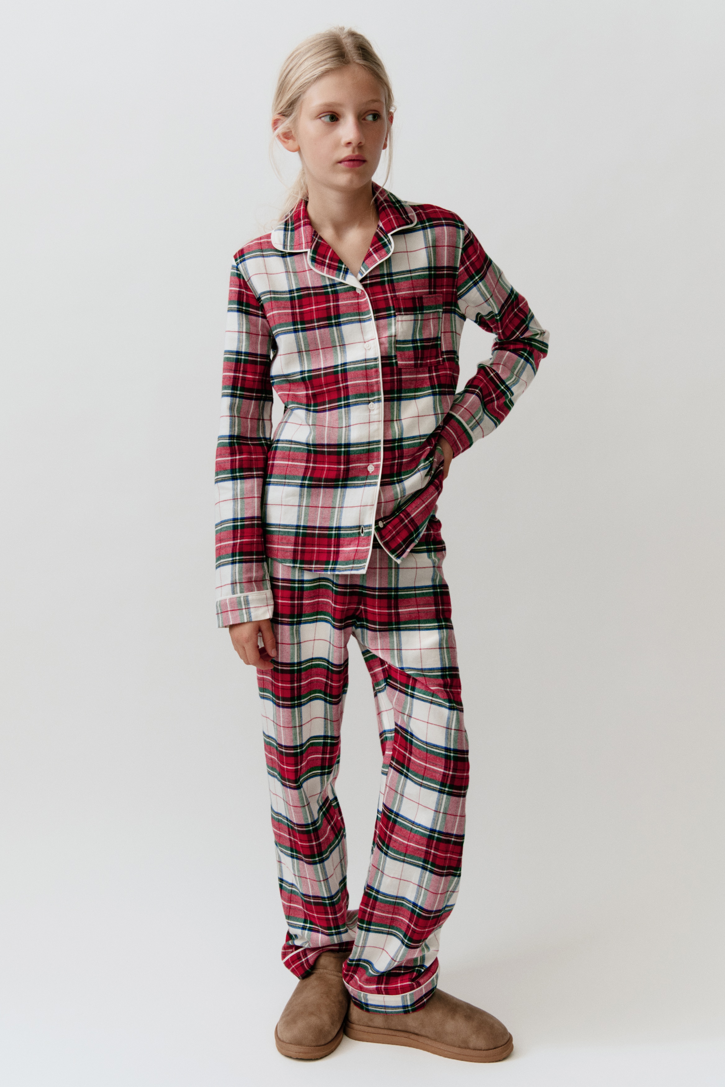 A two-piece flannel pyjama set in a red, white, and dark green plaid pattern is displayed front-facing. The set includes a long-sleeved, button-down top featuring a notch collar, a single chest pocket, and white piping trim, paired with matching straight-leg trousers.