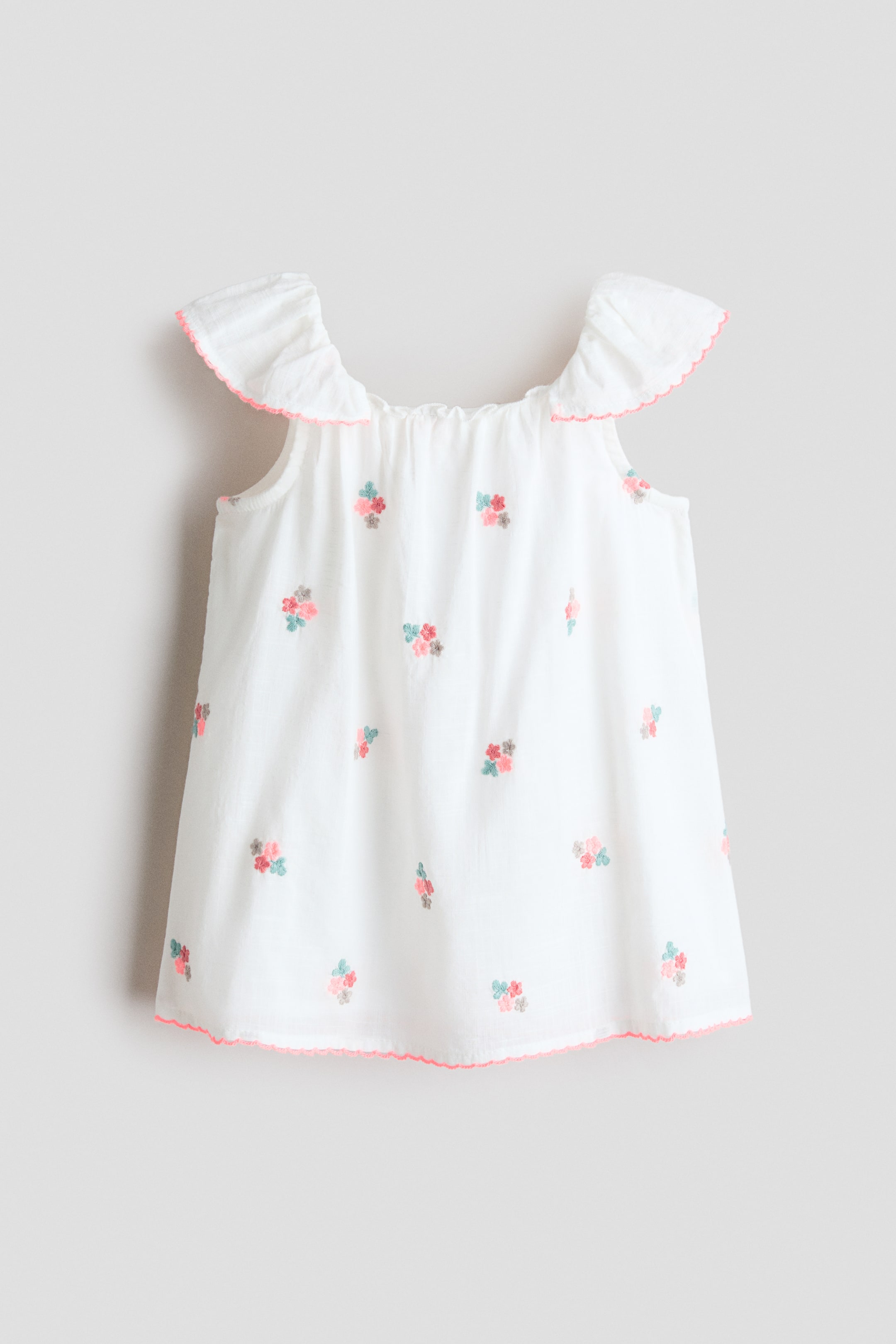 View larger image: Front-facing, the white top showcases a gathered neckline and ruffled cap sleeves with pink scalloped trim. It is embellished with scattered floral embroidery in pink, teal, and grey, and is completed with matching pink scalloped trim along the hem.
