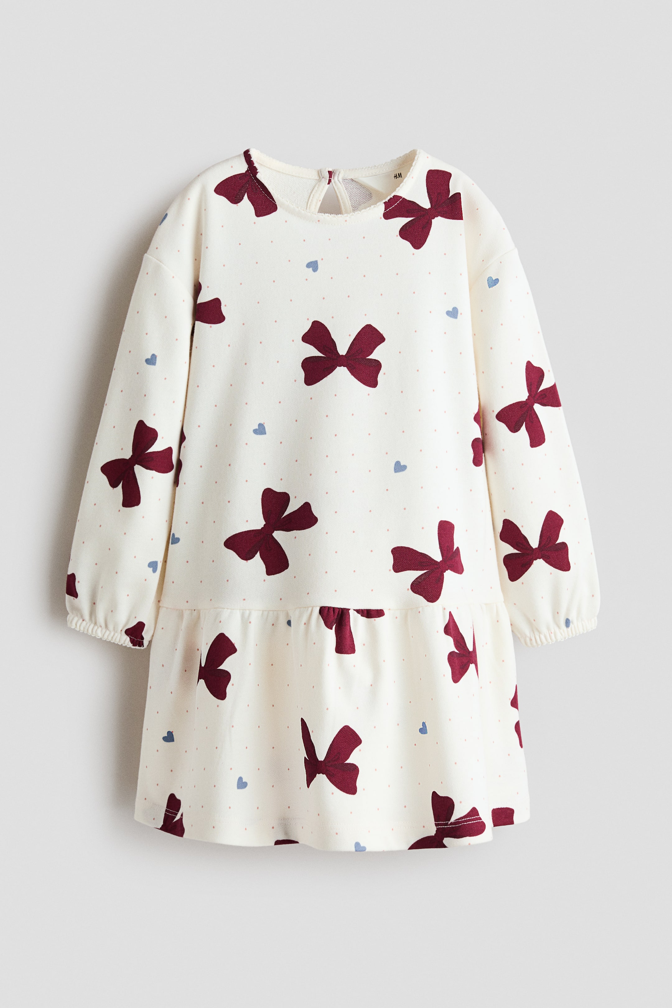 View larger image: A long-sleeved, drop-waist dress is shown facing front, featuring an all-over pattern of burgundy bows, small blue hearts, and light pink dots on an off-white fabric. The dress features a crew neckline with a back keyhole fastening, long sleeves with elasticated cuffs, and a gathered seam flowing into a flared skirt.
