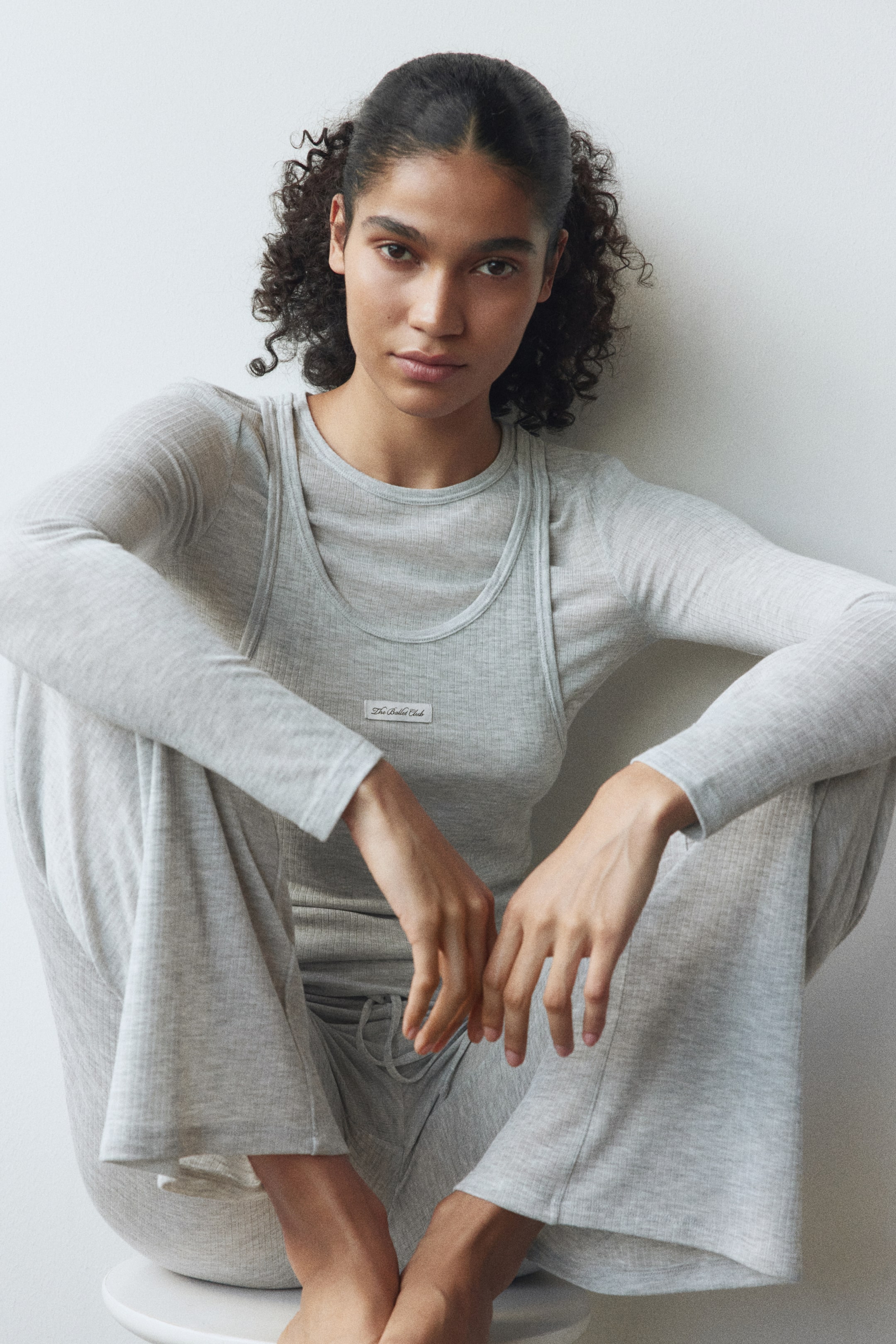 View larger image: The model is wearing a grey ribbed loungewear set. A long-sleeve crew neck top forms the base layer, with a scoop neck tank top layered over it, featuring a small rectangular label on the chest, while wide-leg pants with a visible drawstring waist complete the ensemble.