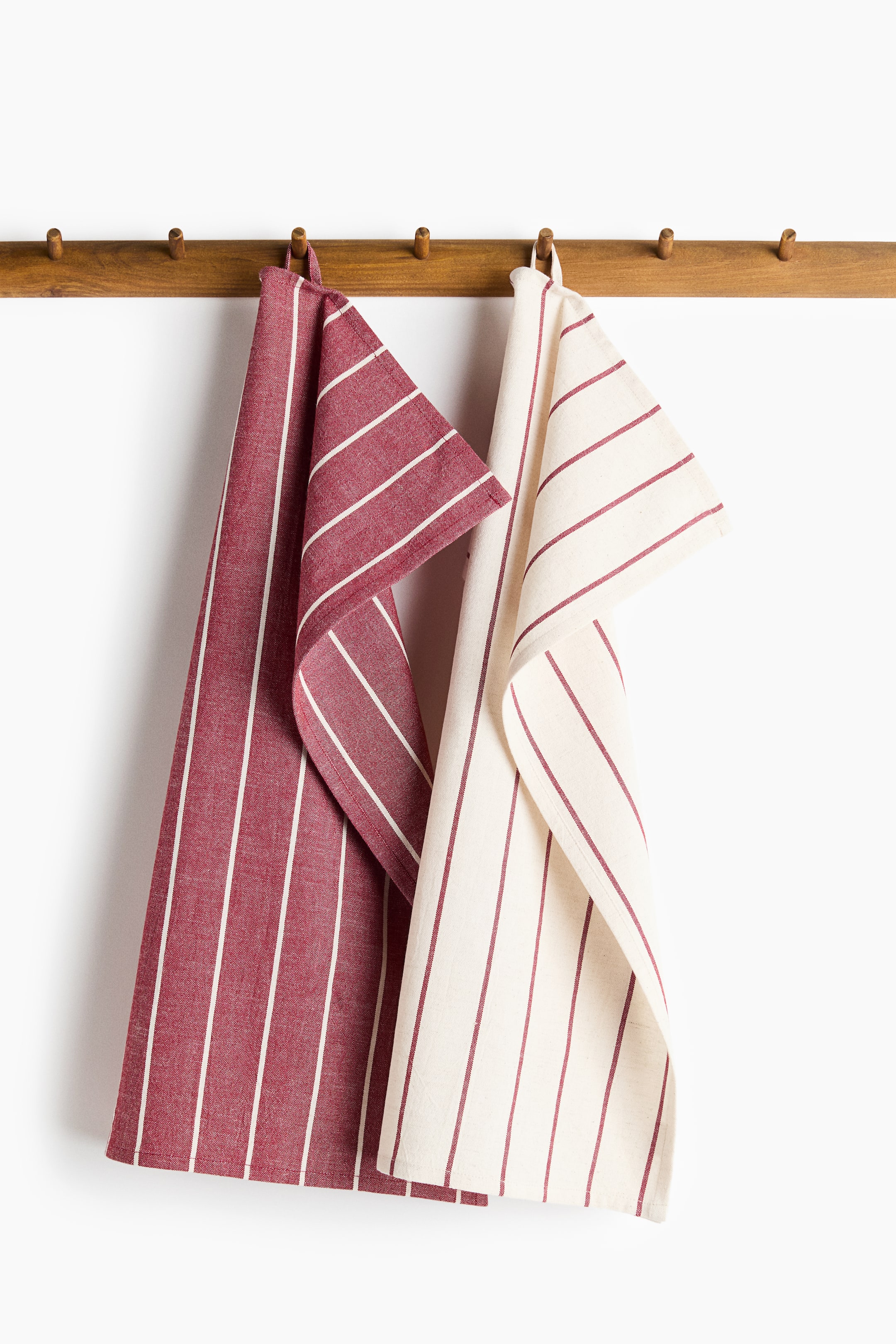 Voir une image plus grande: Deux torchons en mélange de coton et de lin sont suspendus à une patère en bois à l’aide de petites boucles en tissu situées sur leurs côtés courts. Le torchon de gauche présente une base rouge foncé avec de fines rayures verticales blanches, tandis que celui de droite présente une base blanc cassé avec de fines rayures verticales rouge foncé.