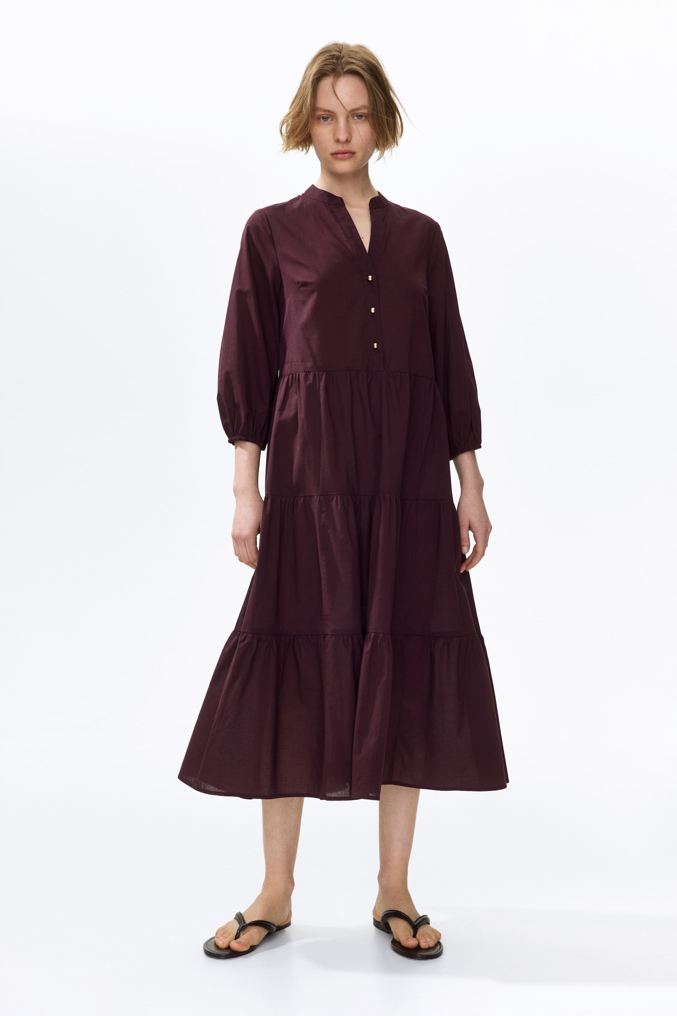 View larger image: The model is wearing a calf-length dark red dress. It boasts a band collar, a V-shaped opening adorned with gold-toned buttons, 3/4-length balloon sleeves with narrow cuffs, and a flared skirt with gathered tiers for added volume.