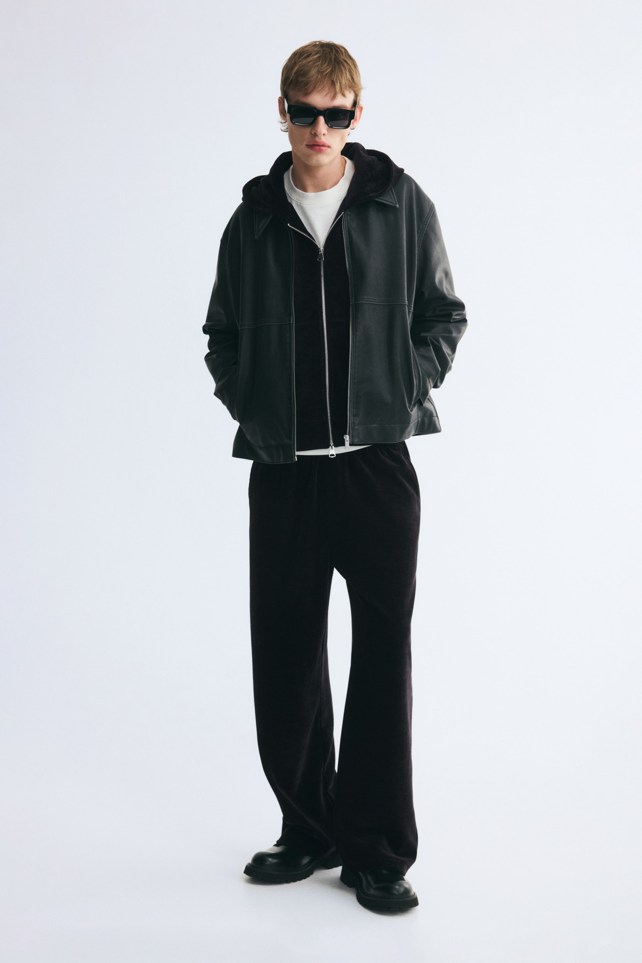 View larger image: A black leather jacket with a classic collar is worn open by the model, who is standing with hands in the side pockets. The jacket is layered over a black zip-up hoodie and a white T-shirt, paired with dark wide-leg trousers.