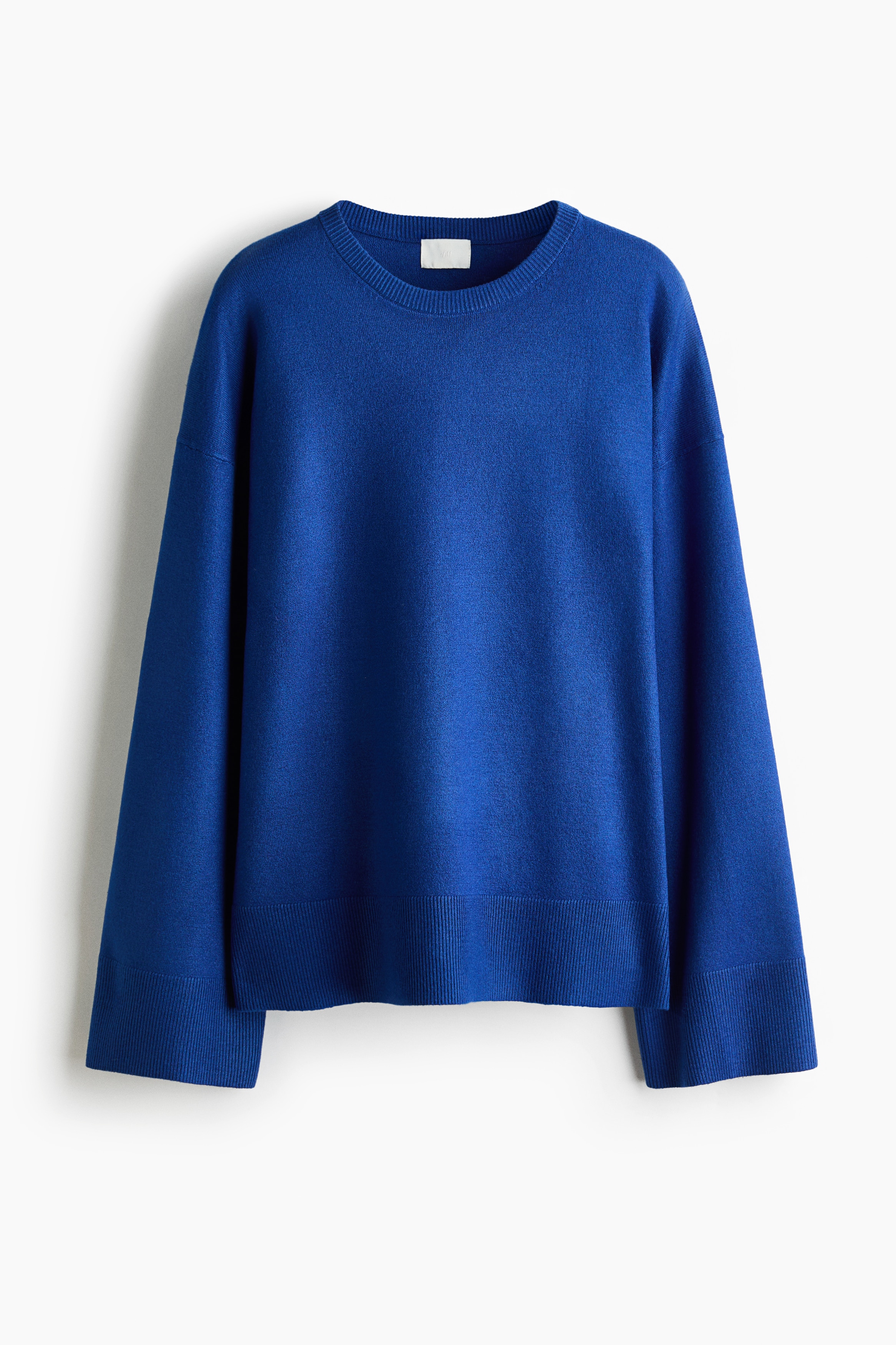 An oversized bright blue knit sweater is shown from the front, featuring dropped shoulders, long sleeves with wide ribbed cuffs, and a ribbed hem with side slits. The sweater is layered over a red and white shirt and styled with a matching blue sweater draped over the shoulders.