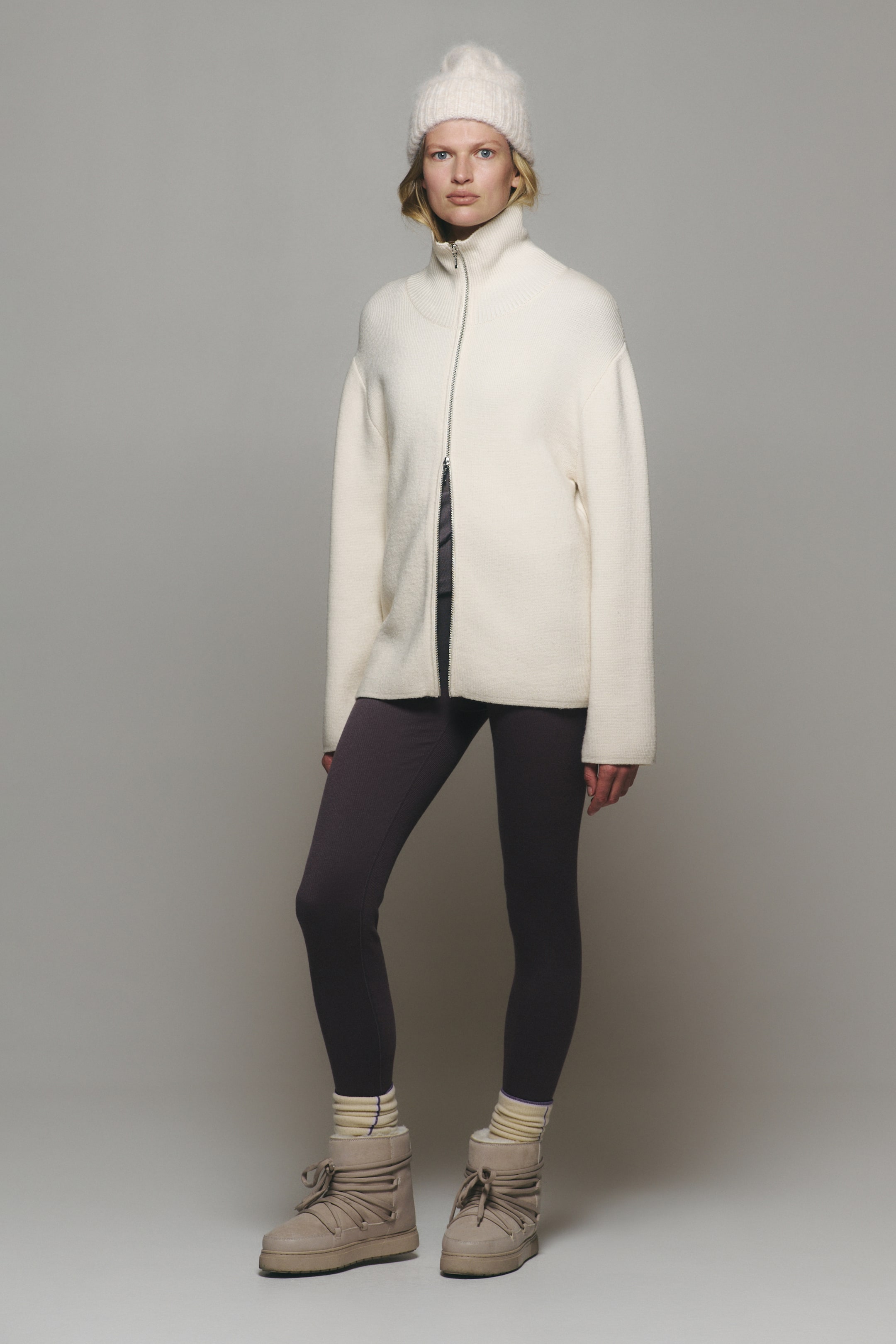 View larger image: An off-white, zip-up knitted jacket featuring a high ribbed collar and long sleeves is being worn by the model. The jacket is styled with dark purple leggings, a matching cream knitted beanie, and tan lace-up winter boots.