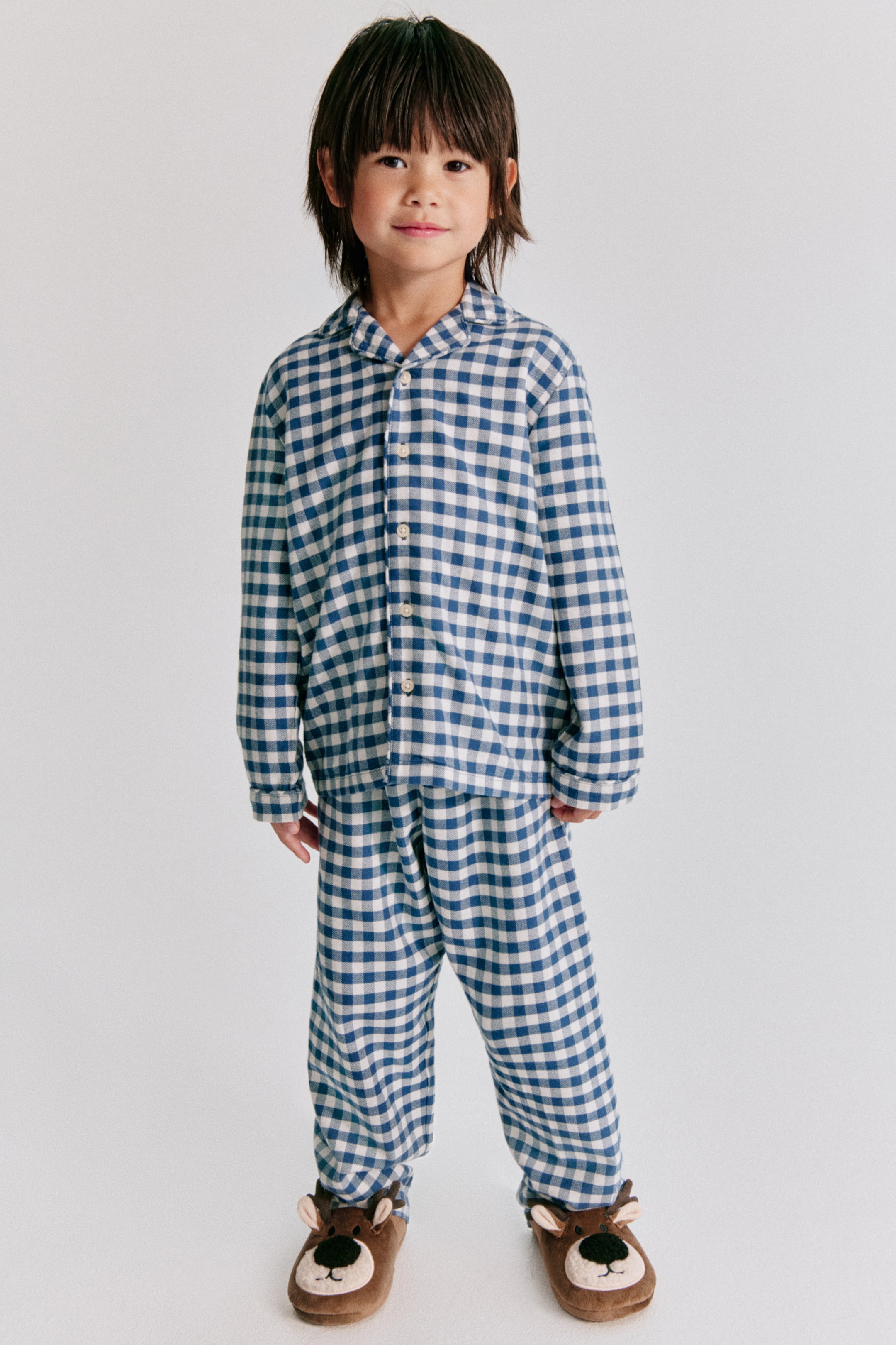 Vezi imaginea mai mare: Un copil poartă o pijama din flanel de bumbac cu model vichy albastru și alb, fotografiat din față pentru a evidenția croiala lejeră. Cămașa cu mânecă lungă are guler, închidere cu nasturi în față și detalii cu paspoal, fiind asortată cu pantaloni drepți în aceeași țesătură.