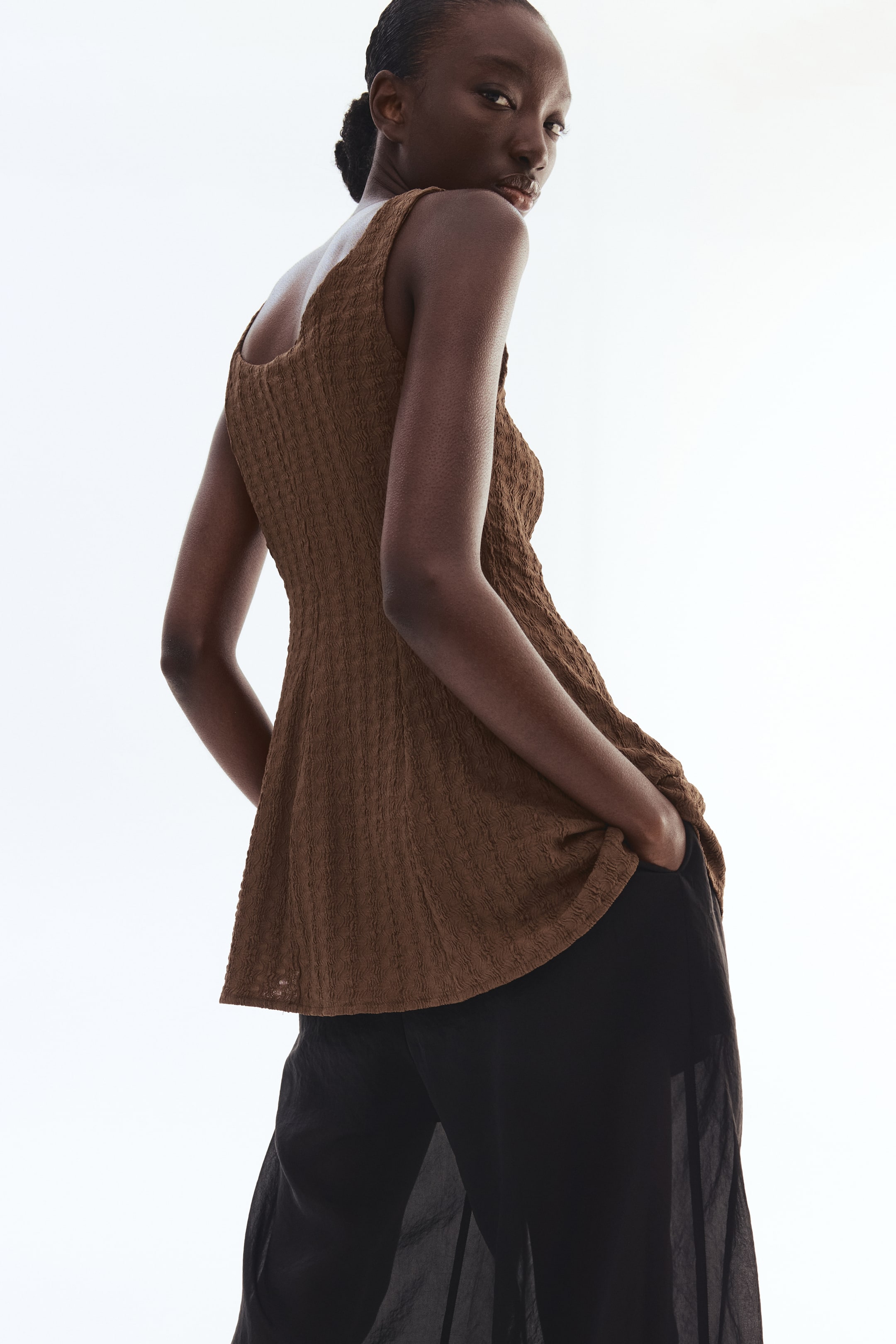 Xem ảnh lớn hơn: Một chiếc áo jersey dài màu nâu có kết cấu, với cổ áo tròn, dây vai rộng và phần peplum xòe, đang được người mẫu mặc. Chiếc áo kéo dài qua quần đen, và tay của người mẫu đang để trong túi quần.