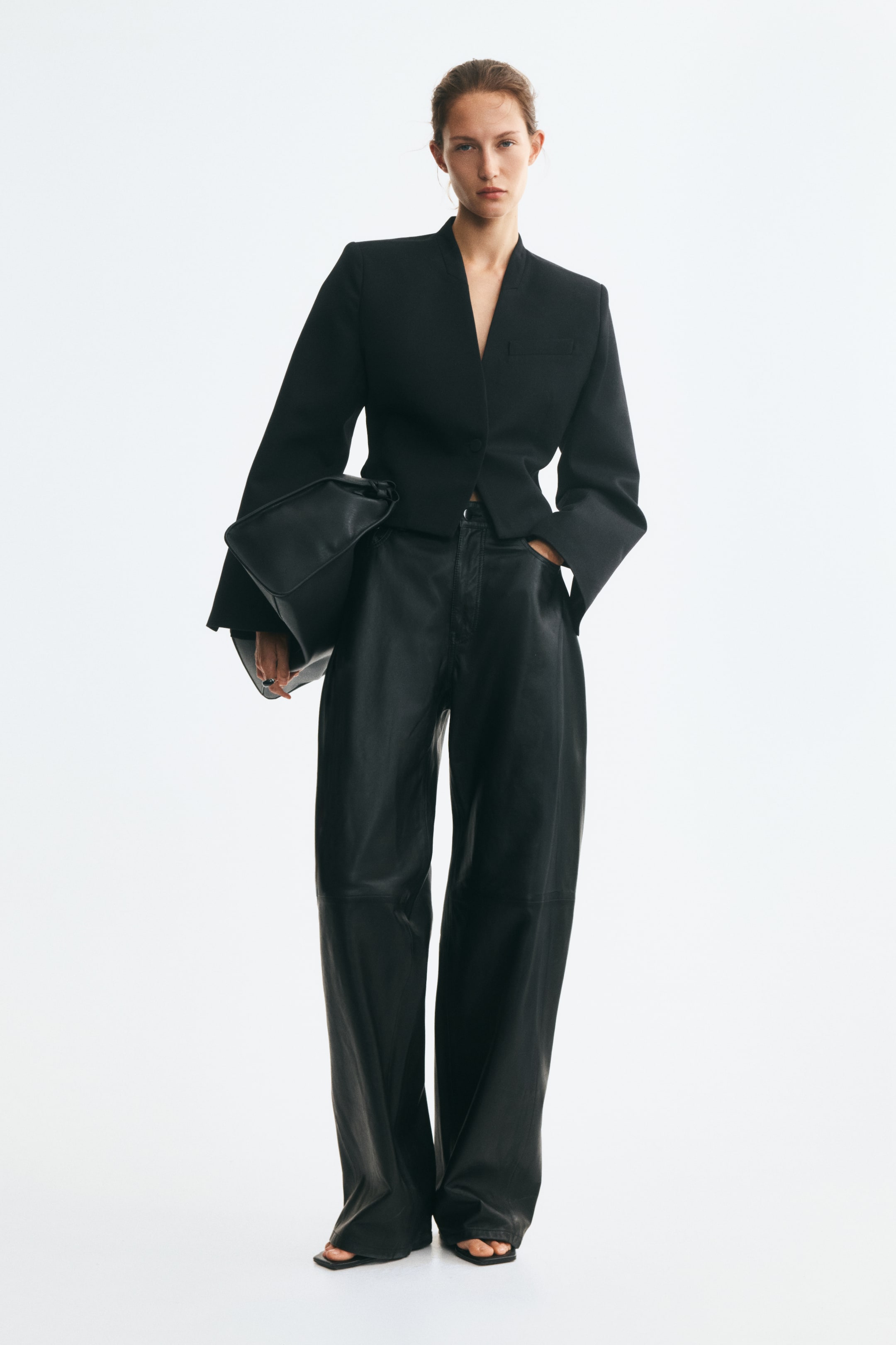 Agrandir l'image: Un pantalon en cuir noir à la coupe ample, taille normale et jambes barrel, avec des coutures horizontales apparentes au niveau des genoux, est porté par le mannequin. Celui-ci pose avec une main dans la poche, associé à un blazer noir court et portant un sac en cuir noir assorti.