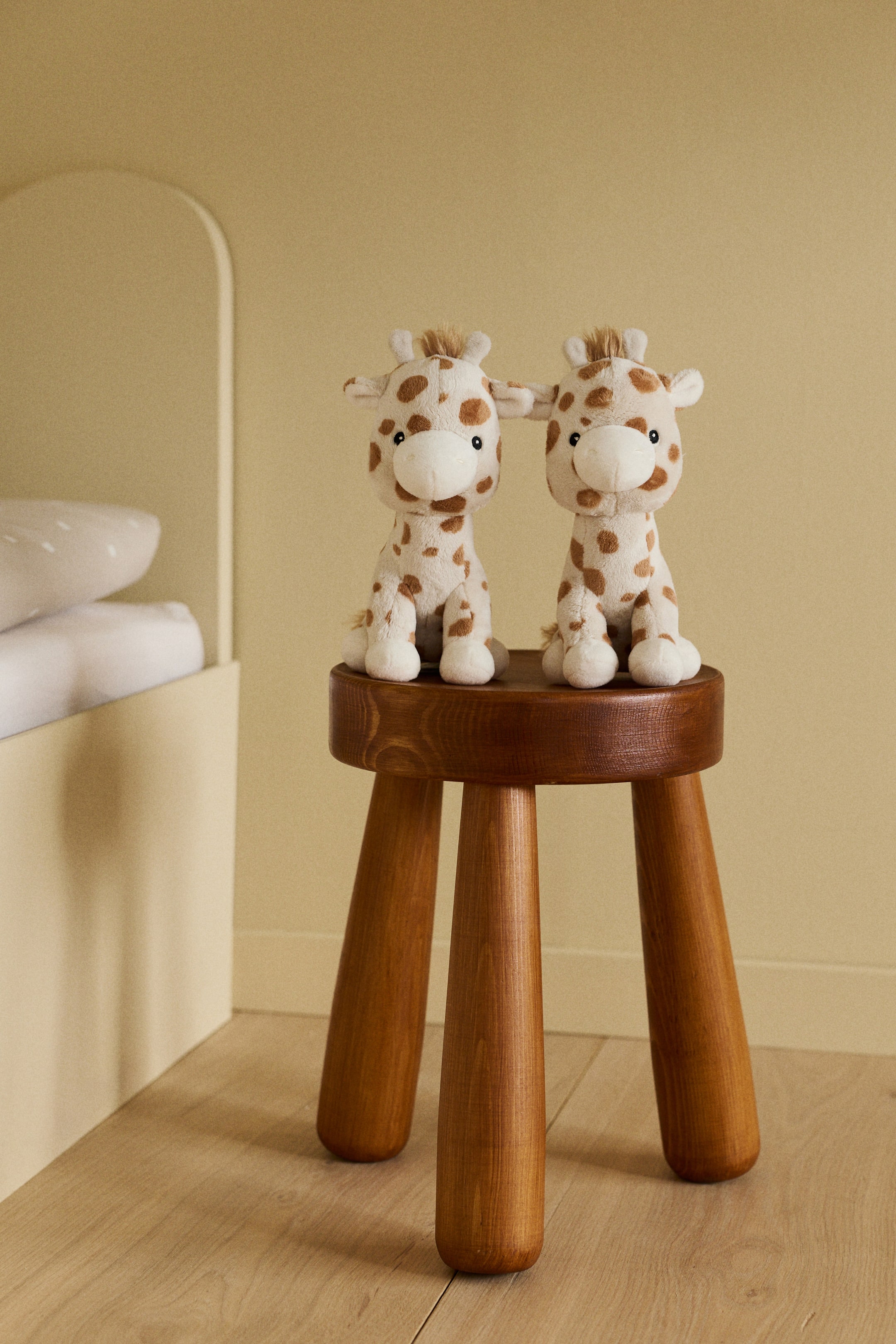 Agrandir l'image: Deux peluches de girafes sont assises côte à côte sur un tabouret en bois rond, faisant face directement vers l'avant. Chaque jouet présente un corps crème avec des taches brunes, un museau blanc, des yeux noirs brodés et de petites cornes touffues.