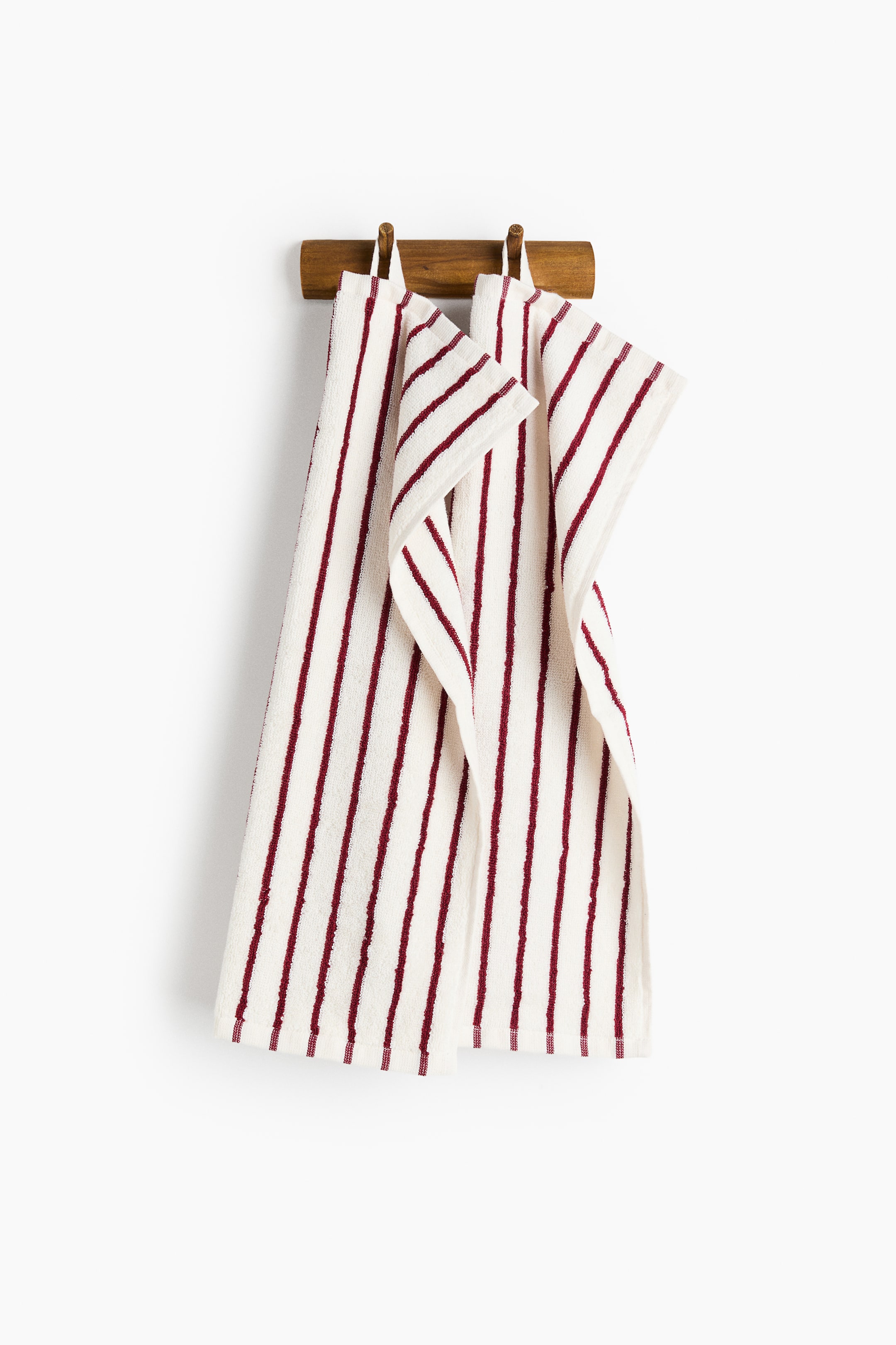 View larger image: A pair of off-white cotton terry guest towels featuring vertical dark red stripes are hanging by their top loops from a wooden peg rail. The towels drape naturally, showcasing their textured fabric, classic striped pattern, and woven hems.