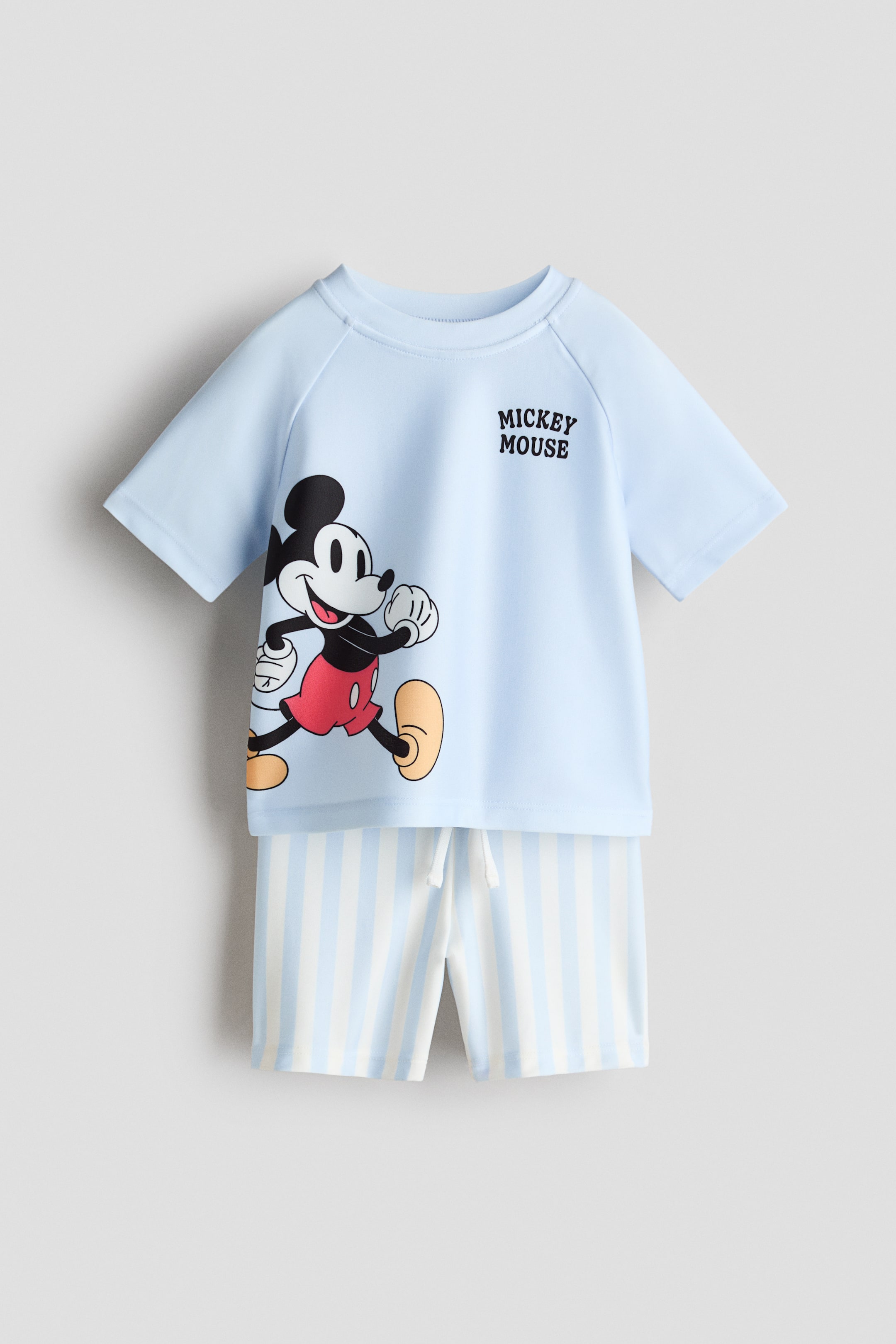 Agrandir l'image: Un ensemble de bain bleu clair, présenté de face, comporte un haut à manches courtes avec un grand visuel de Mickey Mouse et l’inscription «Mickey Mouse». Le haut est doté de manches raglan courtes et d’une encolure ronde, et le short de bain coordonné présente des rayures verticales bleu clair et blanches ainsi qu’un cordon de serrage blanc à la taille.
