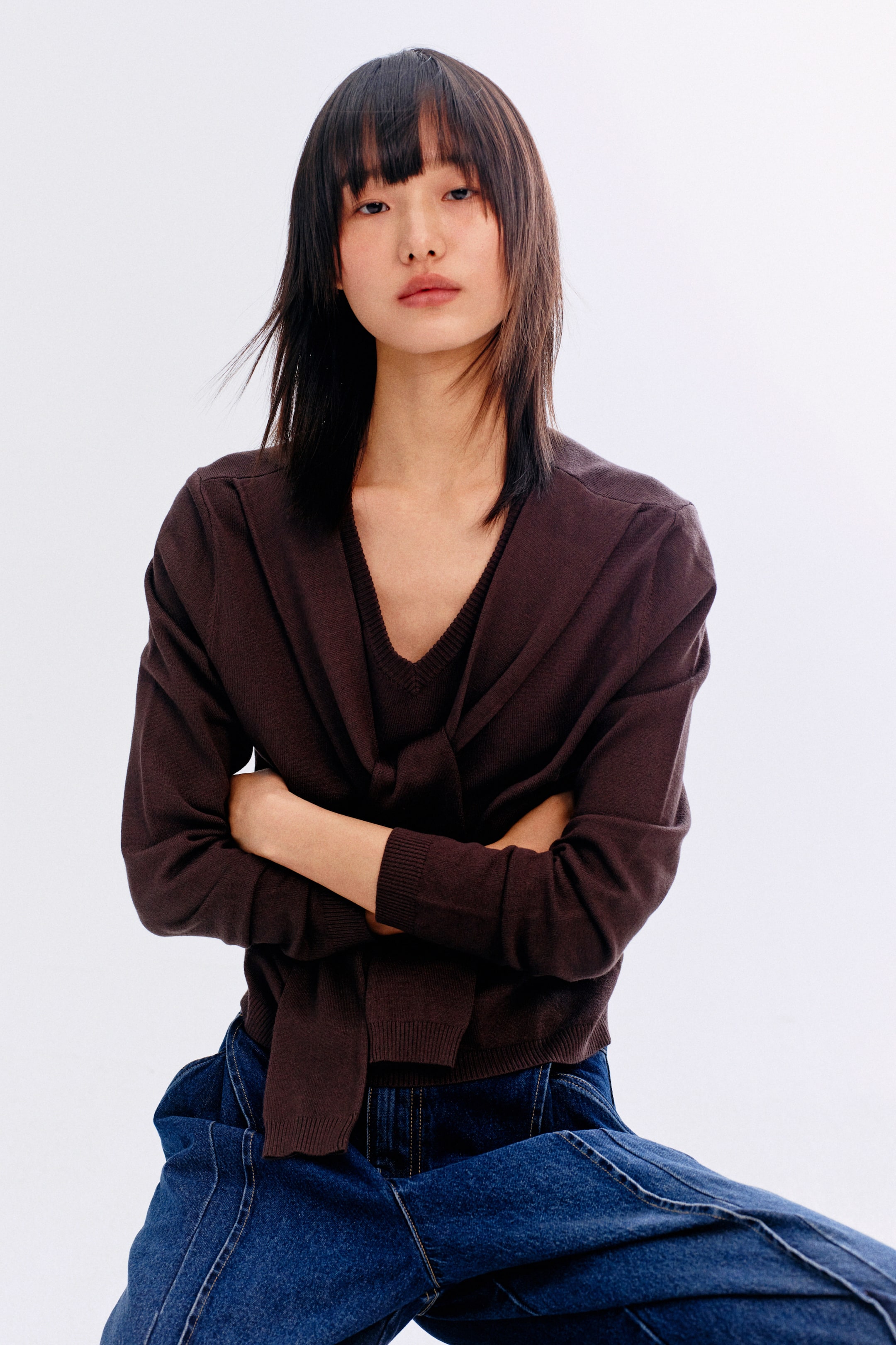 拡大画像を表示: ダークブラウンのニットツインセットが紹介されています。Vネックのトップスとコーディネートされたカーディガンで構成されています。カーディガンは前で結び目を作っており、長袖にはリブカフスとリブ編みの裾が特徴です。