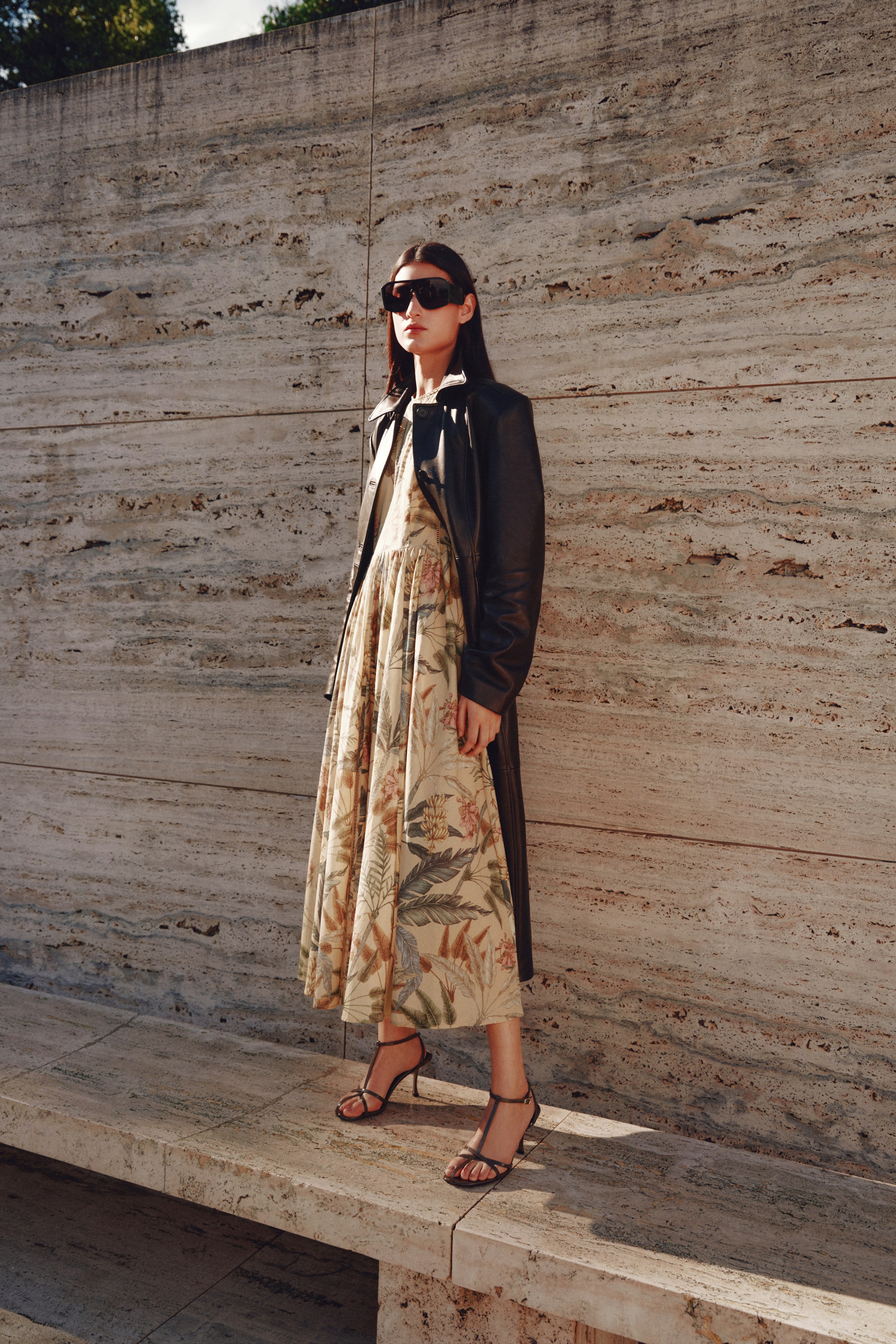 View larger image: A long black leather coat is worn open over a cream midi dress, featuring a full, flowing skirt with an all-over botanical print. Dark strappy high heels and large black sunglasses are also visible, with the entire ensemble presented from a slight front angle.