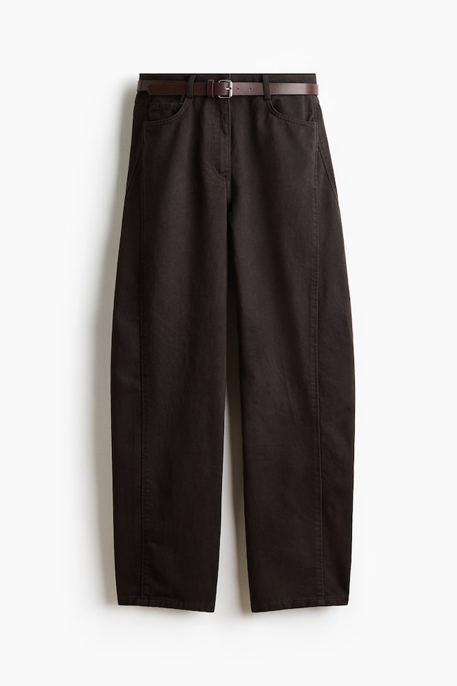Donkerbruine denim jeans met hoge taille, van voren getoond, met een gebogen, volumineuze pijp die naar de zoom toe taps toeloopt. De jeans is gestyled met een smalle, bijpassende bruine riem met zilverkleurige gesp en heeft opvallende gebogen naden langs de zijkanten. - 1