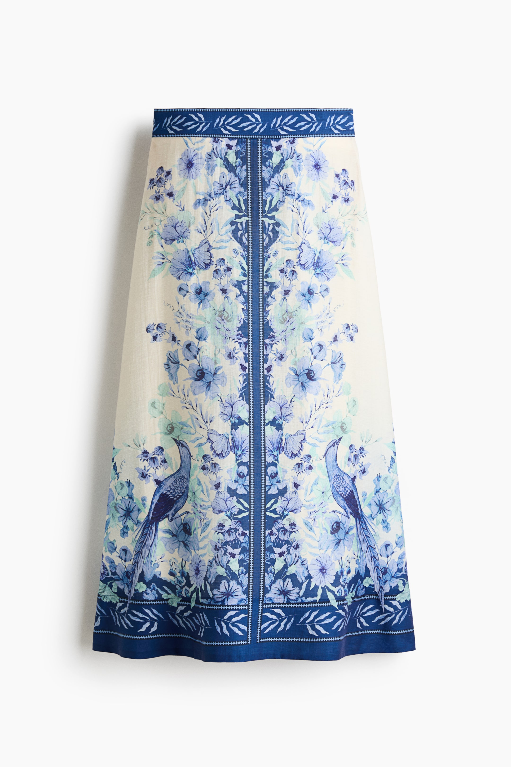 Ampliar la imagen: Una falda de línea A color crema presenta un estampado de flores azules y aves en su parte frontal. Un panel vertical azul oscuro corre por el centro, flanqueado por flores azules más claras y dos pavos reales azules, con bandas azules estampadas a juego que detallan la cintura y el dobladillo.