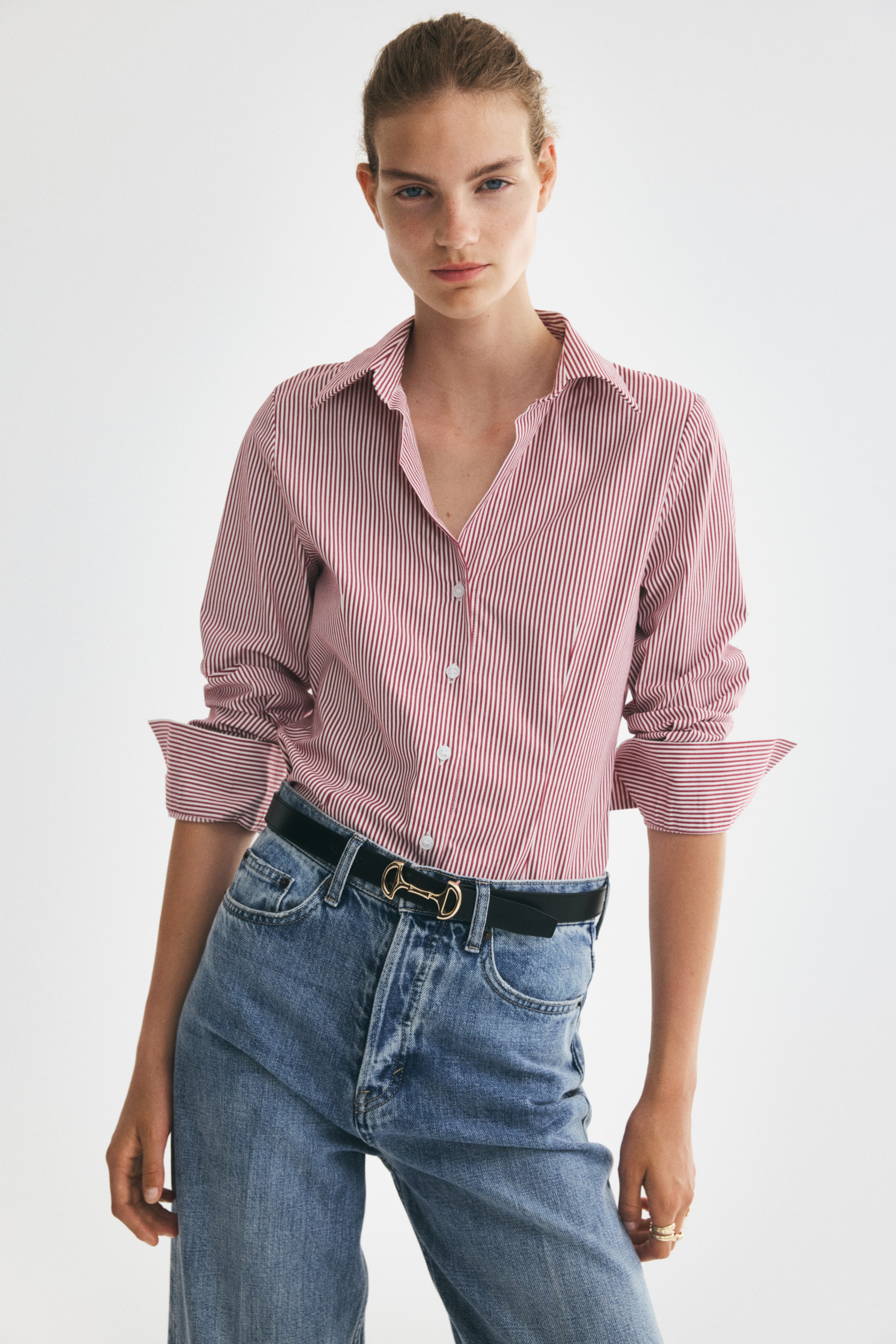 View larger image: The model is wearing a red and white striped fitted shirt. The shirt features an open collar, buttons down the front, and long sleeves that are rolled up. It is tucked into blue jeans, which are secured with a black belt.