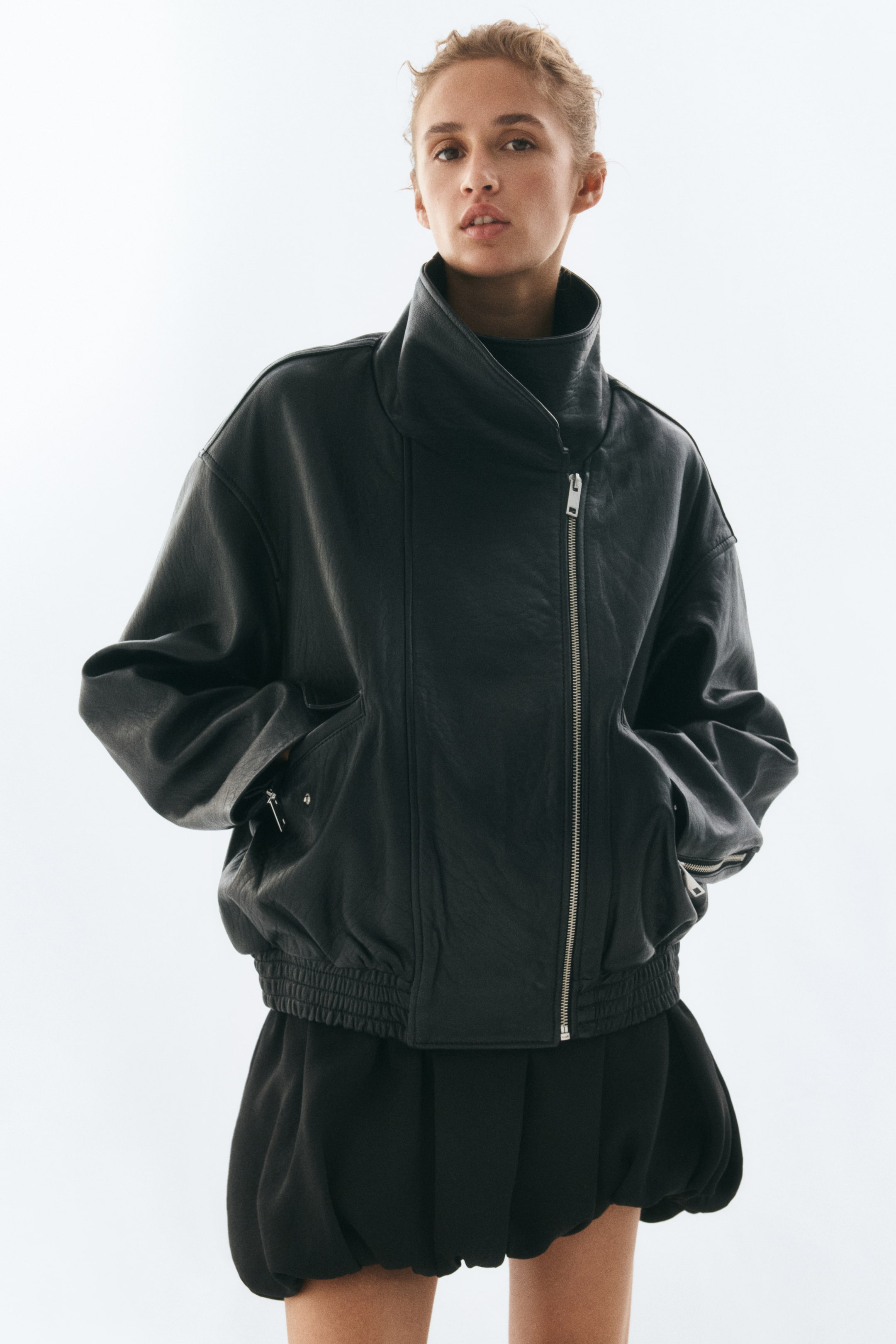 Agrandir l'image: Une veste en cuir noire oversize, dotée d’un col cheminée montant, d’une fermeture zippée asymétrique sur le devant et d’un ourlet smocké élastiqué, est portée par le modèle. Le modèle a les mains placées dans les poches à rabat frontales, et associe la veste à une jupe noire à ourlet boule.