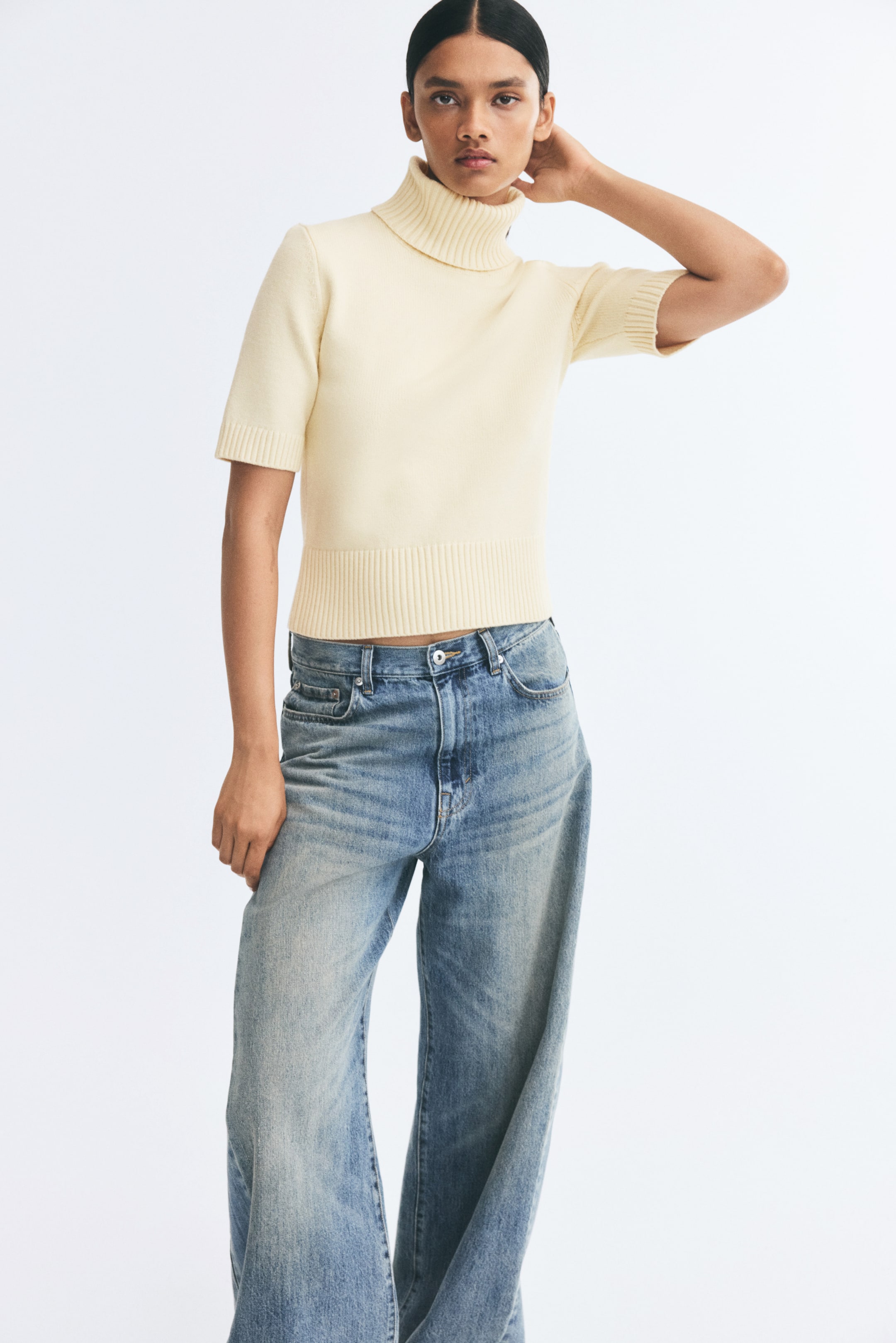 View larger image: The model is wearing a light yellow knitted top featuring a ribbed turtleneck, short sleeves with ribbed cuffs, and a wide ribbed hem. The fitted top is styled with light blue wide-leg jeans.