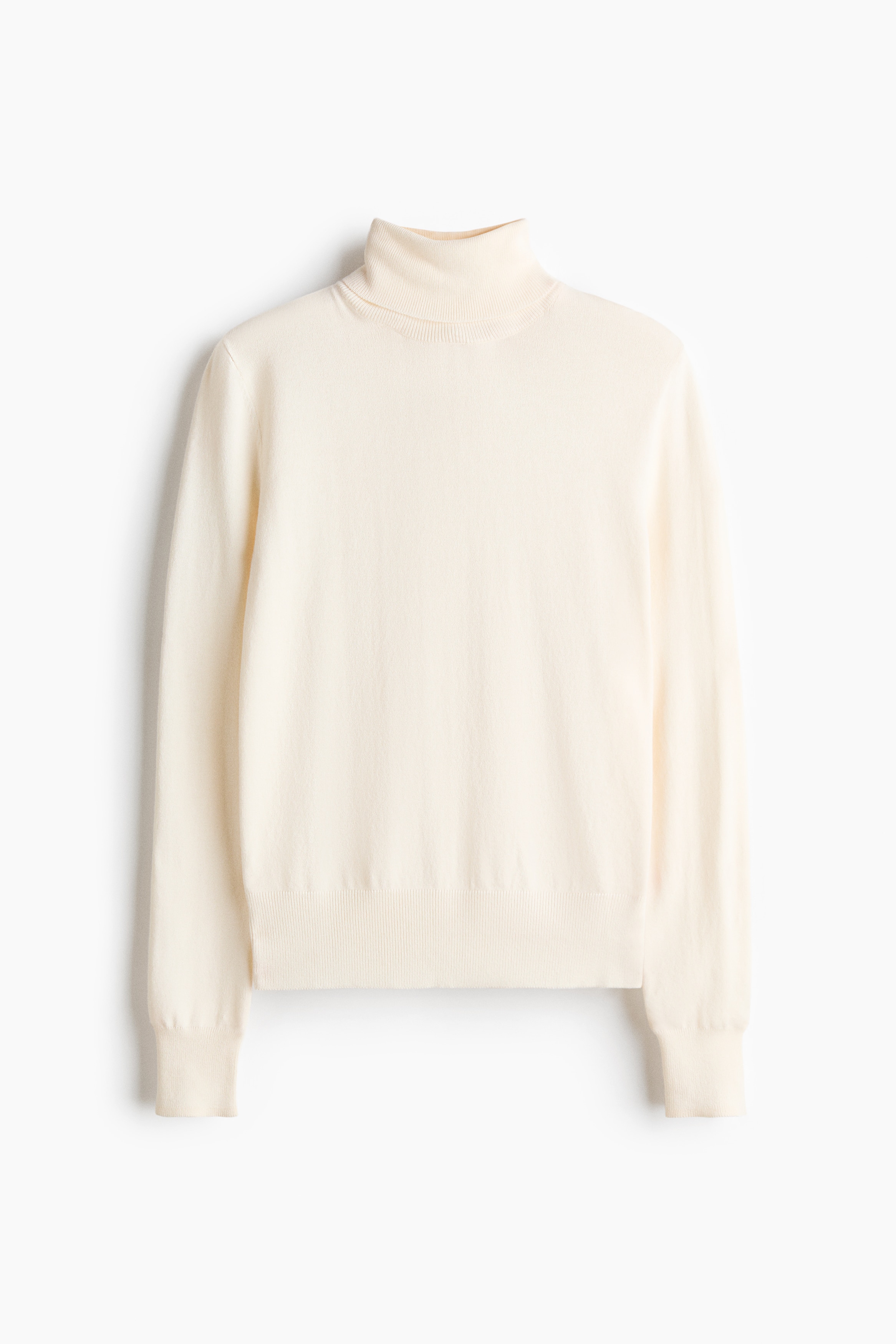 En naturvit finstickad trÃ¶ja med ribbad polokrage, lÃ¥nga Ã¤rmar och ribbad fÃ¥ll bÃ¤rs av modellen. TrÃ¶jan Ã¤r stylad med vita byxor och ett svart bÃ¤lte med ett spÃ¤nne i silverfÃ¤rg.