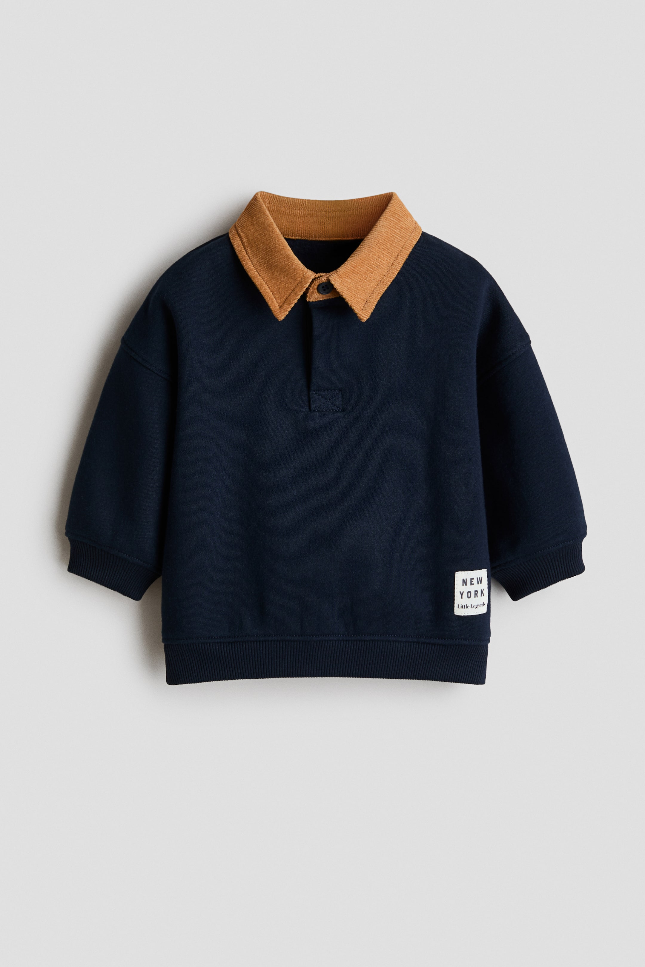 Büyük resmi görüntüle: Lacivert bir sweatshirt, kontrast oluşturan kahverengi fitilli yaka ve ön kısmında iki düğmeli patlet detayına sahiptir. Ayrıca fitilli manşetler, fitilli etek ucu ve ön sağ alt kısmında "NEW YORK Little Legends" yazılı beyaz dikdörtgen bir etiket de bulunmaktadır.