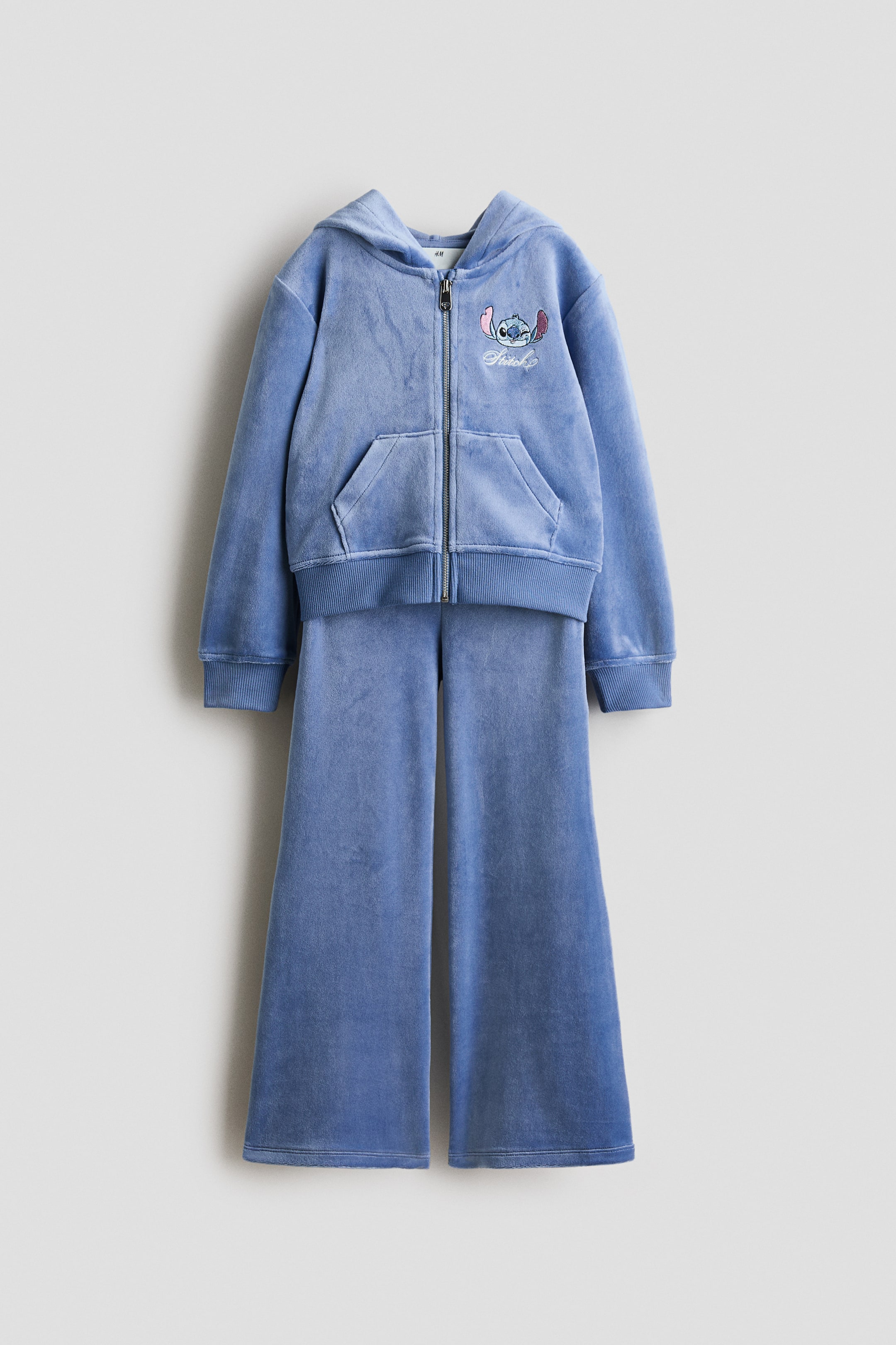 View larger image: A blue velour tracksuit features a full-zip hoodie and wide-leg trousers. The hoodie has a hood, a split kangaroo pocket, ribbed cuffs and a ribbed hem at the front, along with an embroidered Stitch character and "Stitch" text on the left chest. The matching wide-leg trousers complement the hoodie.