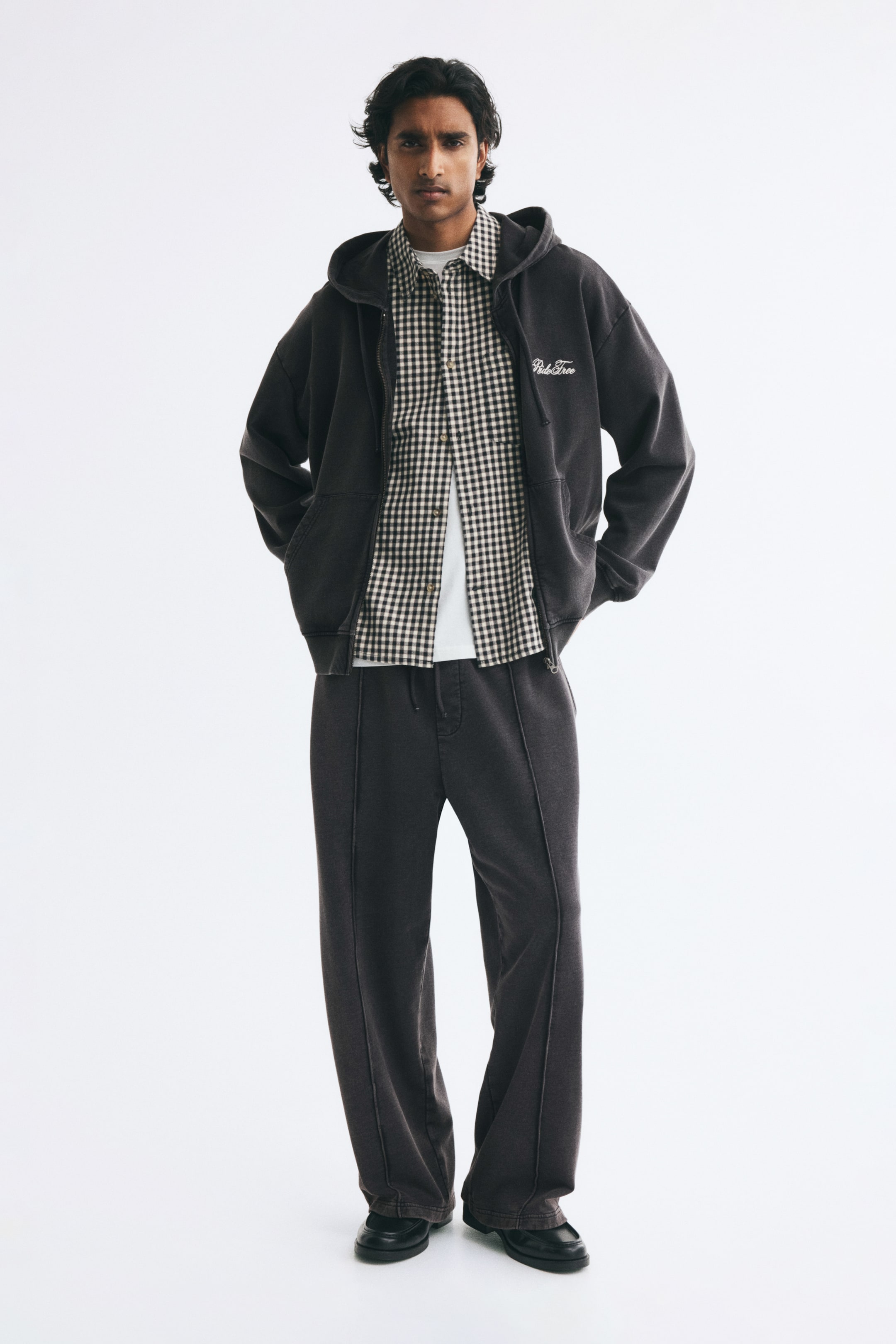 View larger image: The model is wearing a dark grey zip-up hoodie and matching wide-leg sweatpants with centre-seam detailing. The open hoodie, featuring a small white embroidered logo on the chest, is layered over a black-and-white gingham button-down shirt and a plain white T-shirt.