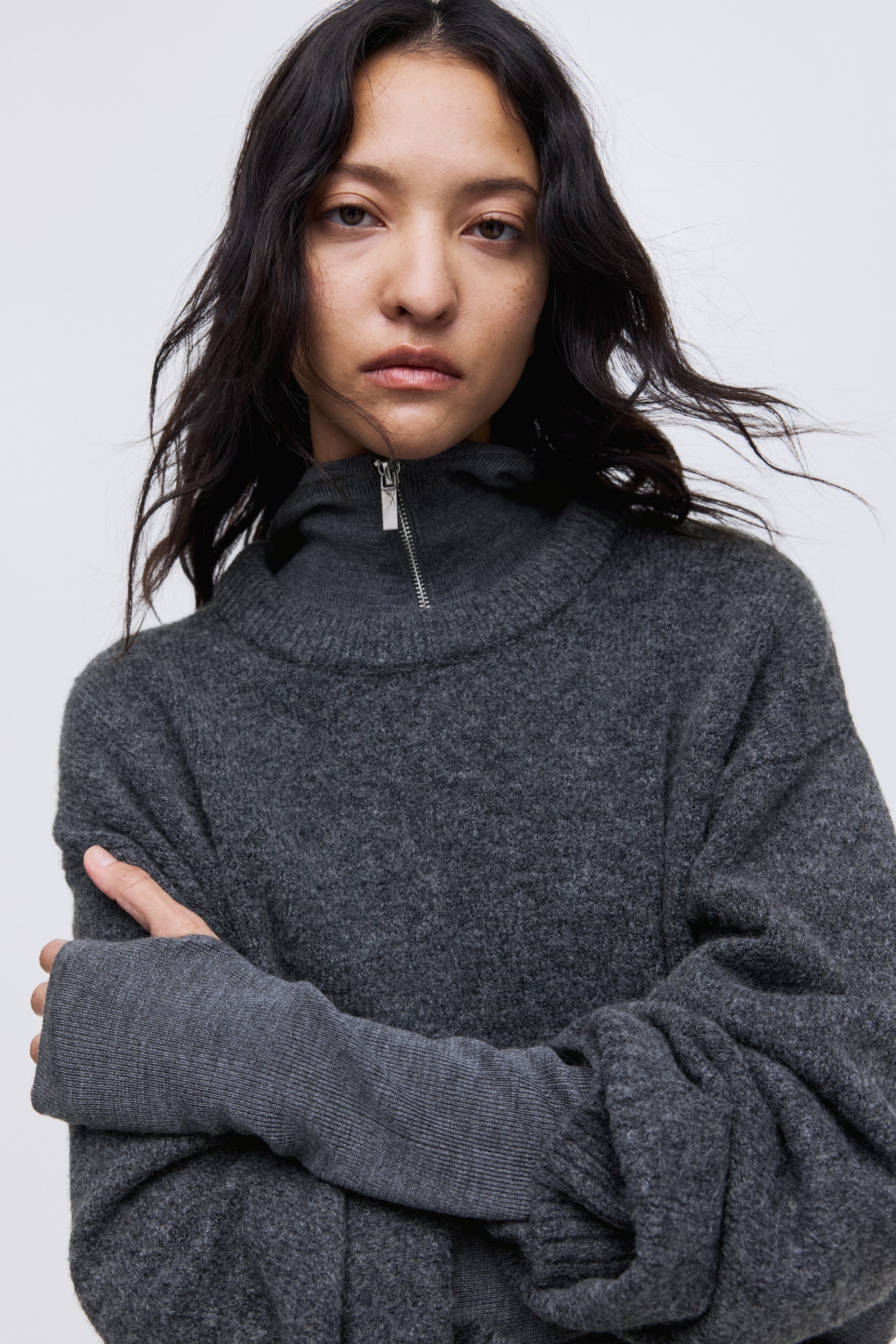 Ver imagen más grande: Un jersey de punto gris oscuro muestra un cuello alto con un cuarto de cremallera en tono plateado parcialmente desabrochado. Sus mangas largas y holgadas terminan con puños acanalados extendidos, uno de los cuales presenta un diseño con agujero para el pulgar.