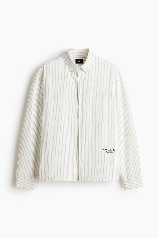 The long-sleeved shirt is showcased from the front, boasting a cream base adorned with subtle dark vertical stripes. It features a button-down collar and a full button-front closure. Additionally, it highlights a left chest patch pocket, buttoned cuffs, and black embroidered text reading 'Cross-Country Champs' on the right chest. - 1