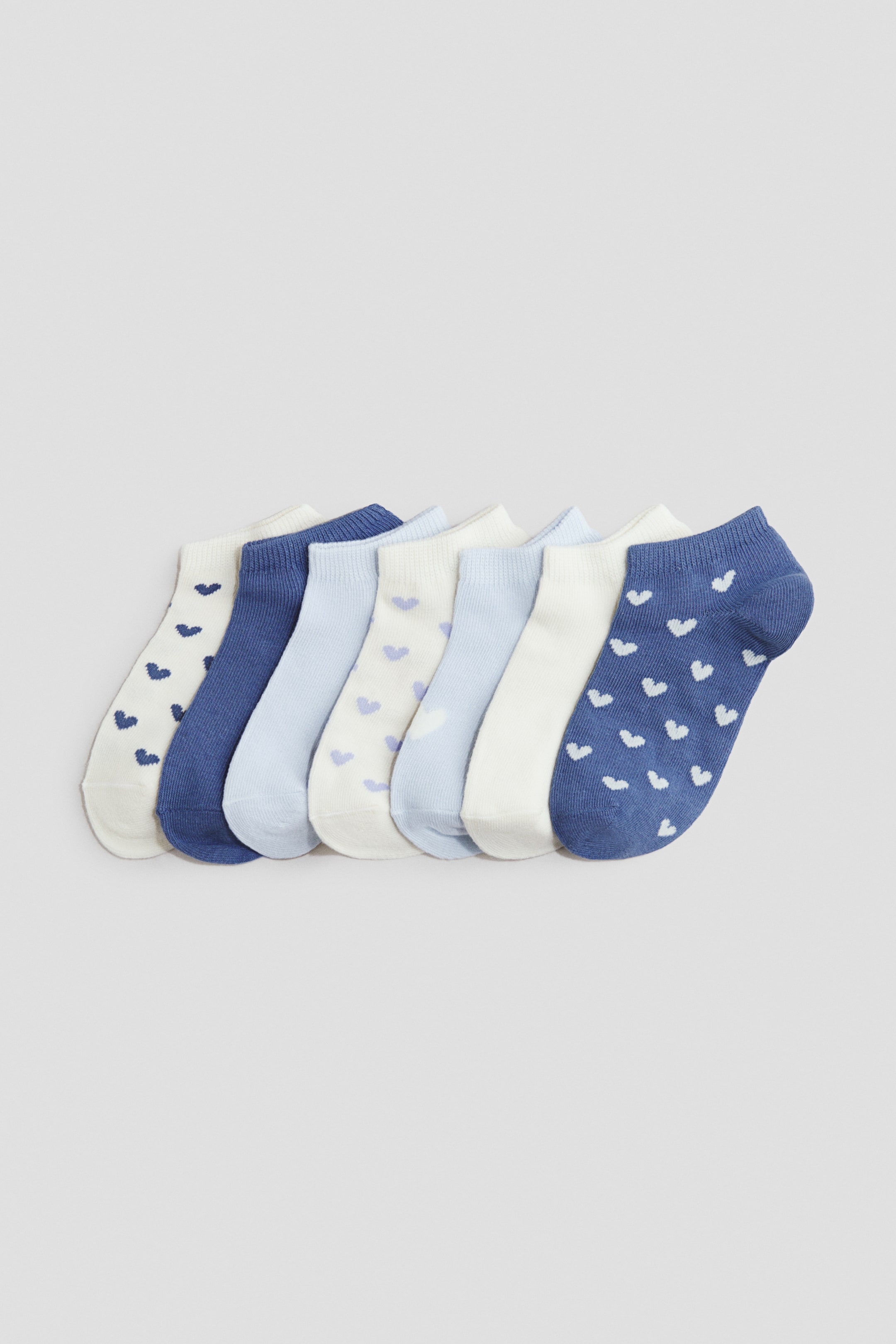 View larger image: "Fine-knit trainer socks are arranged in a row, showcasing a range of blue and off-white hues. Some socks boast solid colours, while others display small heart patterns, with elasticated bands visible around their tops."