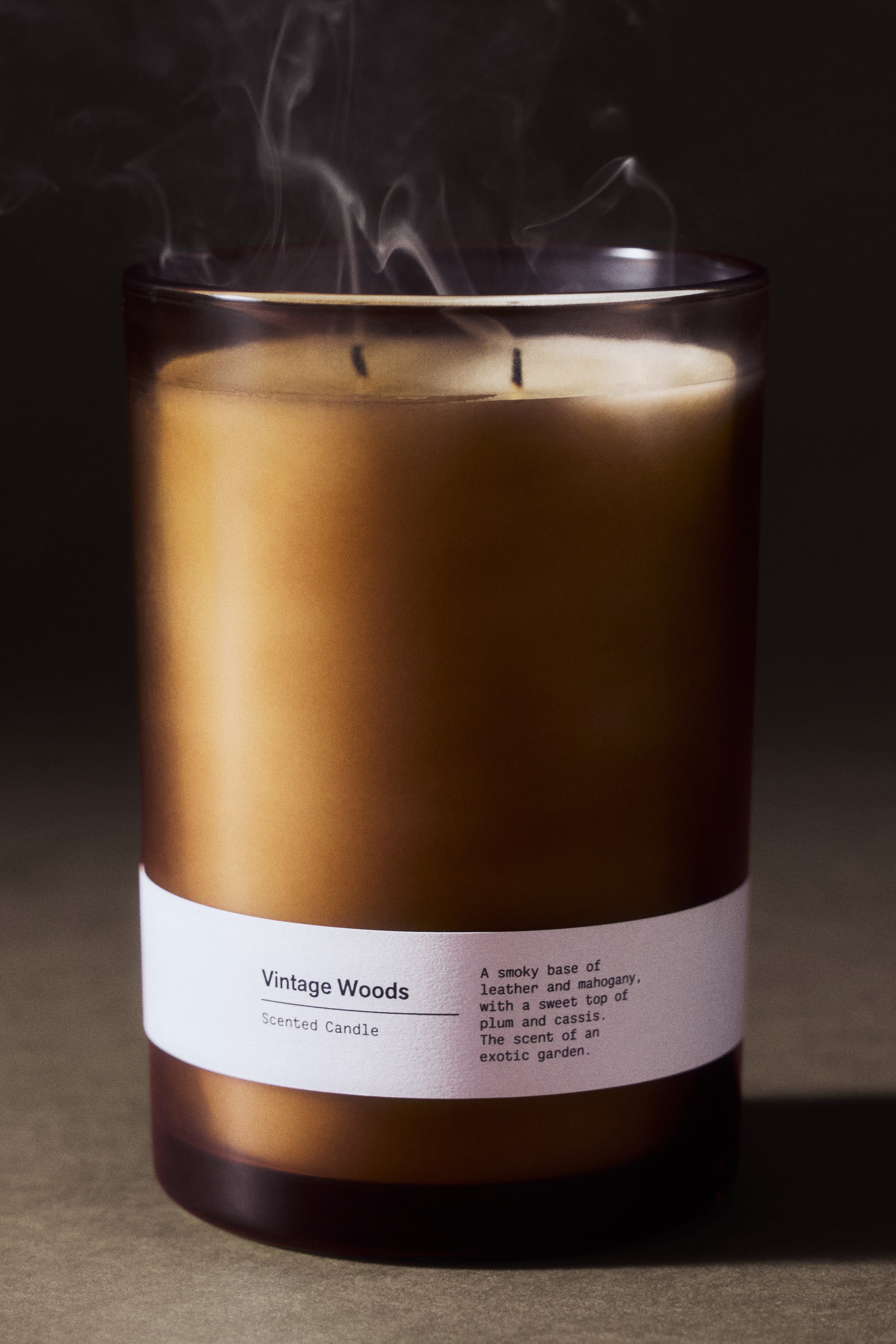 View larger image: A front view of a scented candle in a translucent brown glass holder, featuring a white paper label with the text "Vintage Woods." Thin wisps of smoke are rising from the double wicks set in light golden wax.