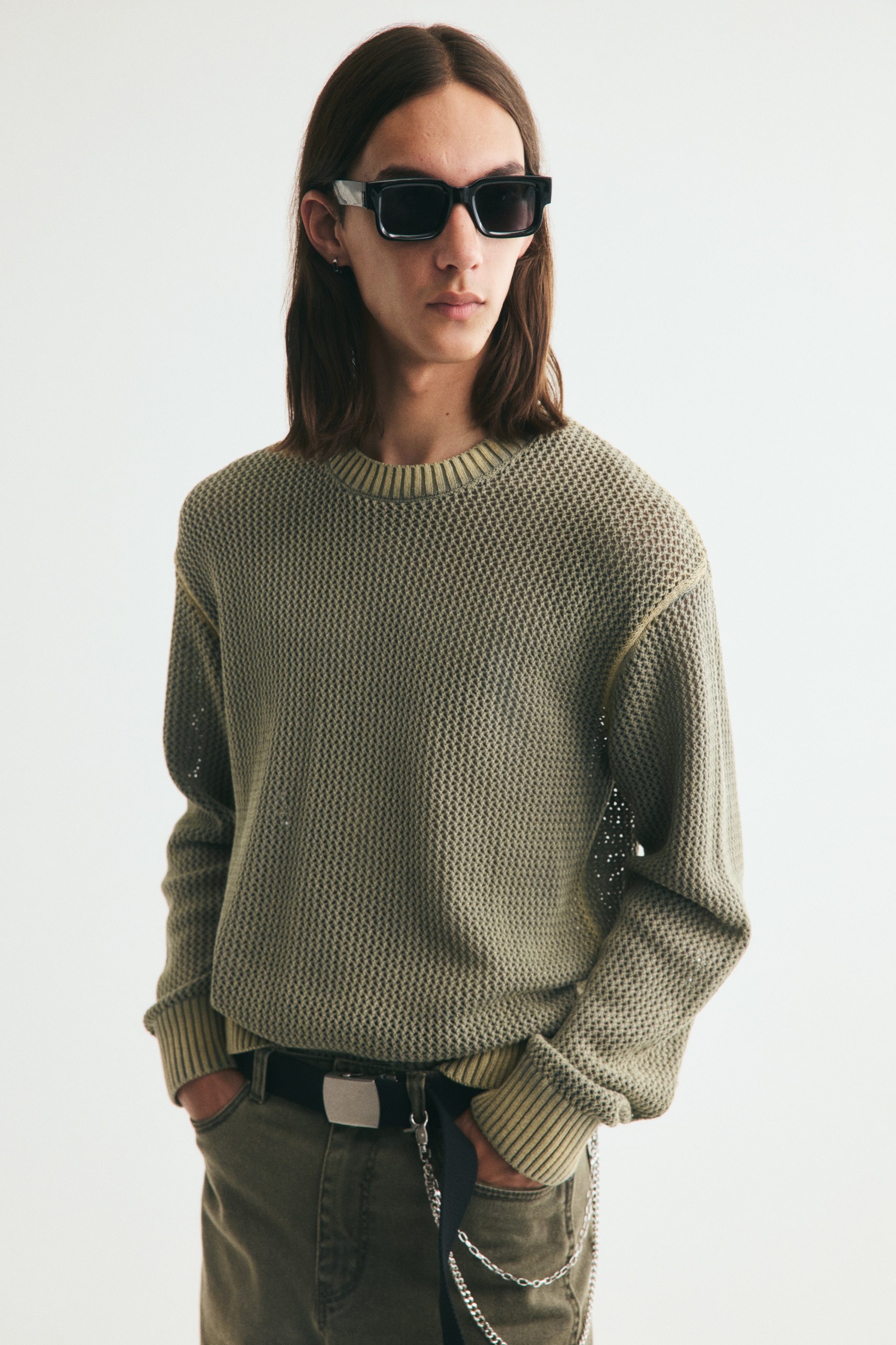 The olive green open-knit sweater is shown from the front, showcasing its textured design. It features a ribbed crew neck, ribbed cuffs, and a ribbed hem, all in a faded beige hue, which also accentuates the raglan shoulder seams.