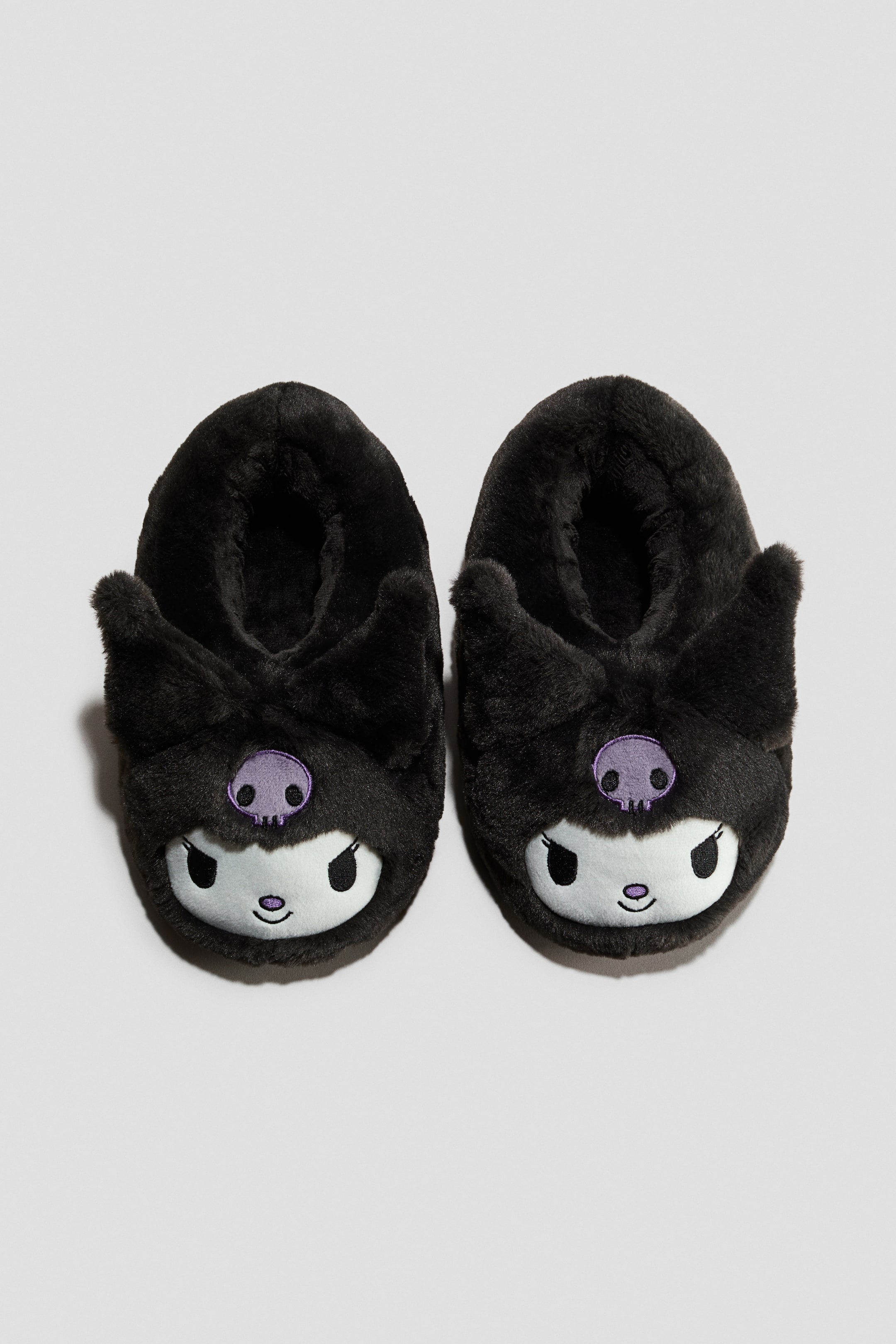 Grösseres Bild ansehen: Ein Paar schwarze, flauschige Hausschuhe mit einem Charakterdesign auf der Vorderseite; jeder Schuh zeigt ein weisses Gesicht, schwarz gestickte Augen und einen kleinen Mund. Auf der Stirn zwischen zwei spitzen, aufrechten Ohren befindet sich ein violettes Totenkopf-Emblem.