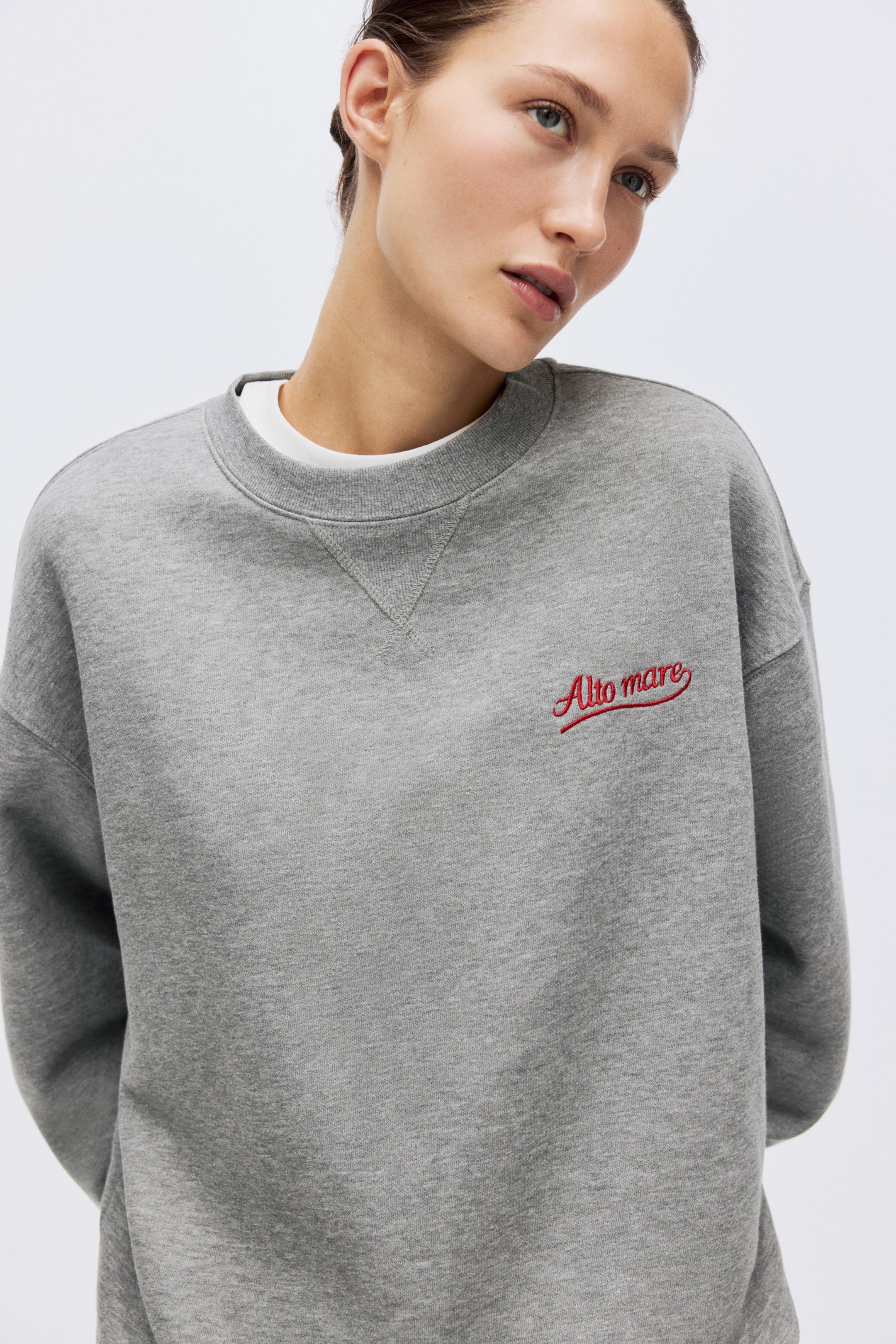View larger image: A grey crew-neck sweatshirt features a ribbed collar with a V-stitch detail below it. Red embroidered text "Alto mare" appears on the left chest, and a white T-shirt is layered beneath the neckline.