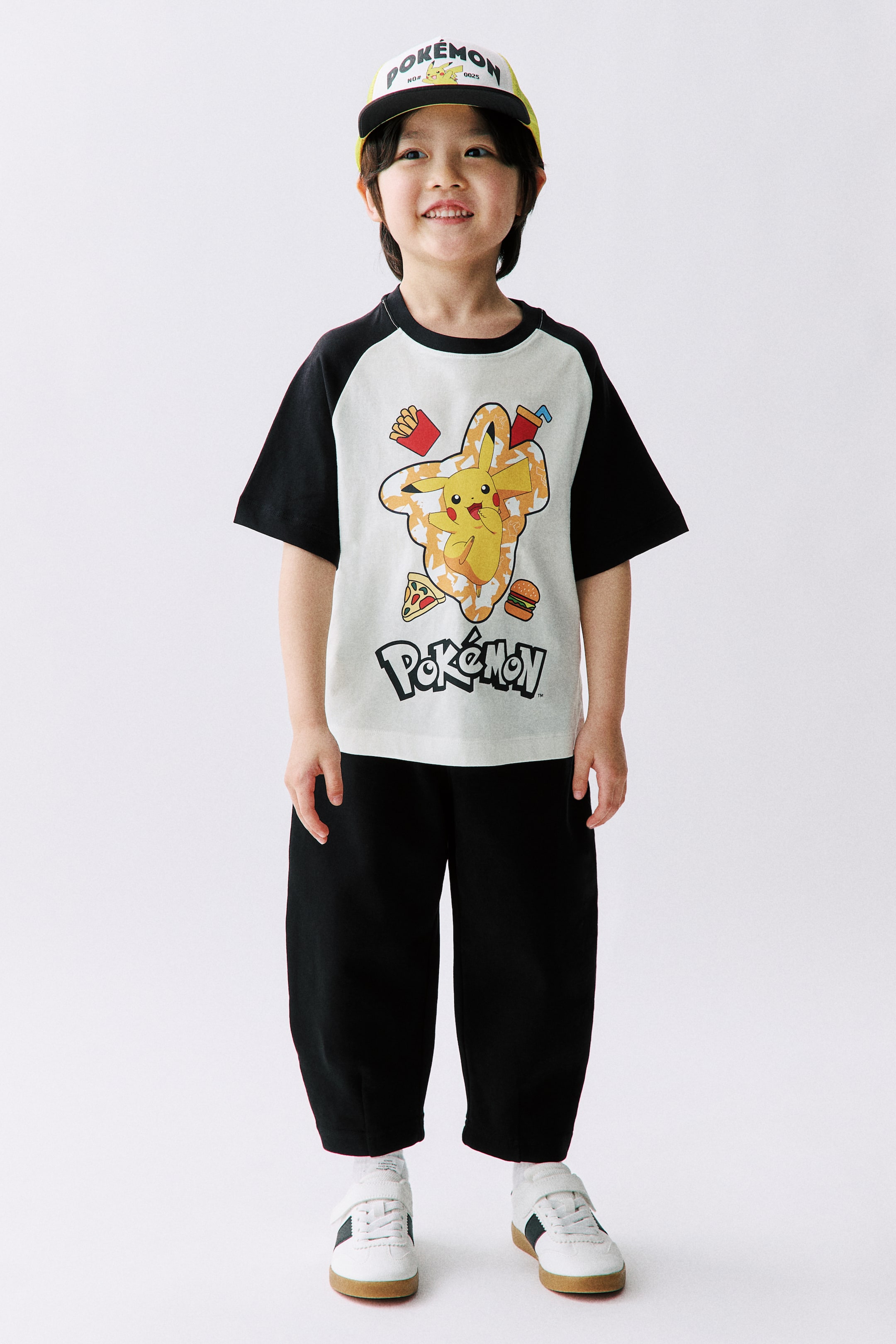 Xem ảnh lớn hơn: Một chiếc mũ bóng chày Pokémon màu trắng và vàng đang được đội, với một dải màu đen phía trên vành mũ màu vàng và hình ảnh Pikachu kèm chữ "Pokémon" ở mặt trước. Một chiếc áo thun raglan màu trắng ngà nhạt với tay áo màu đen đang được mặc, hiển thị hình ảnh Pikachu đầy màu sắc và đồ ăn nhanh ở phía trước. Quần ống rộng màu đen cắt ngắn đang được mặc, kết hợp với giày thể thao màu trắng có sọc bên tối và khóa dán.