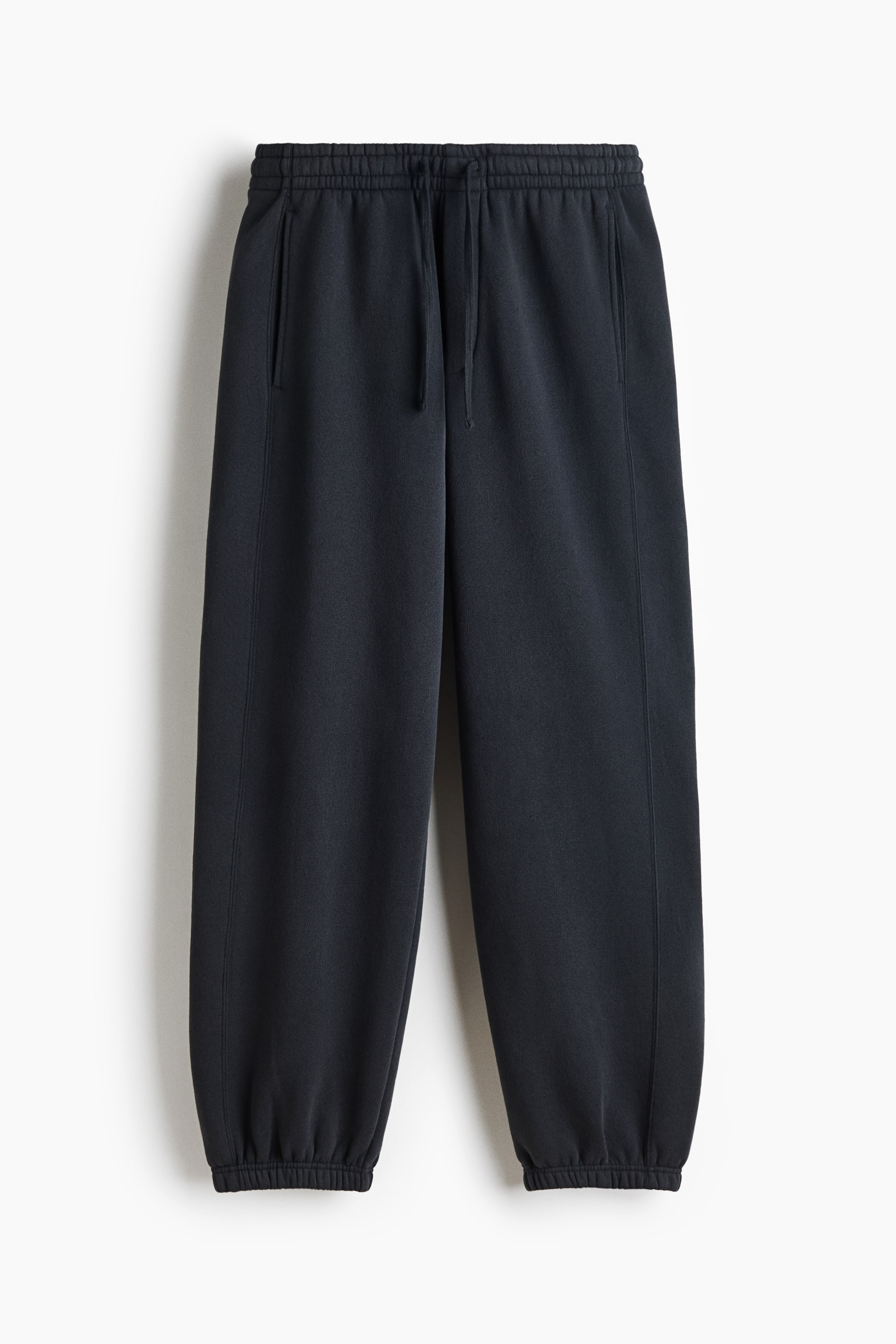 Voir une image plus grande: Une paire de pantalons de survêtement gris foncé est suspendue de face, avec une taille élastiquée munie d’un cordon de serrage assorti. Le jogger coupe ample présente des poches latérales verticales et des poignets élastiques resserrés aux chevilles.