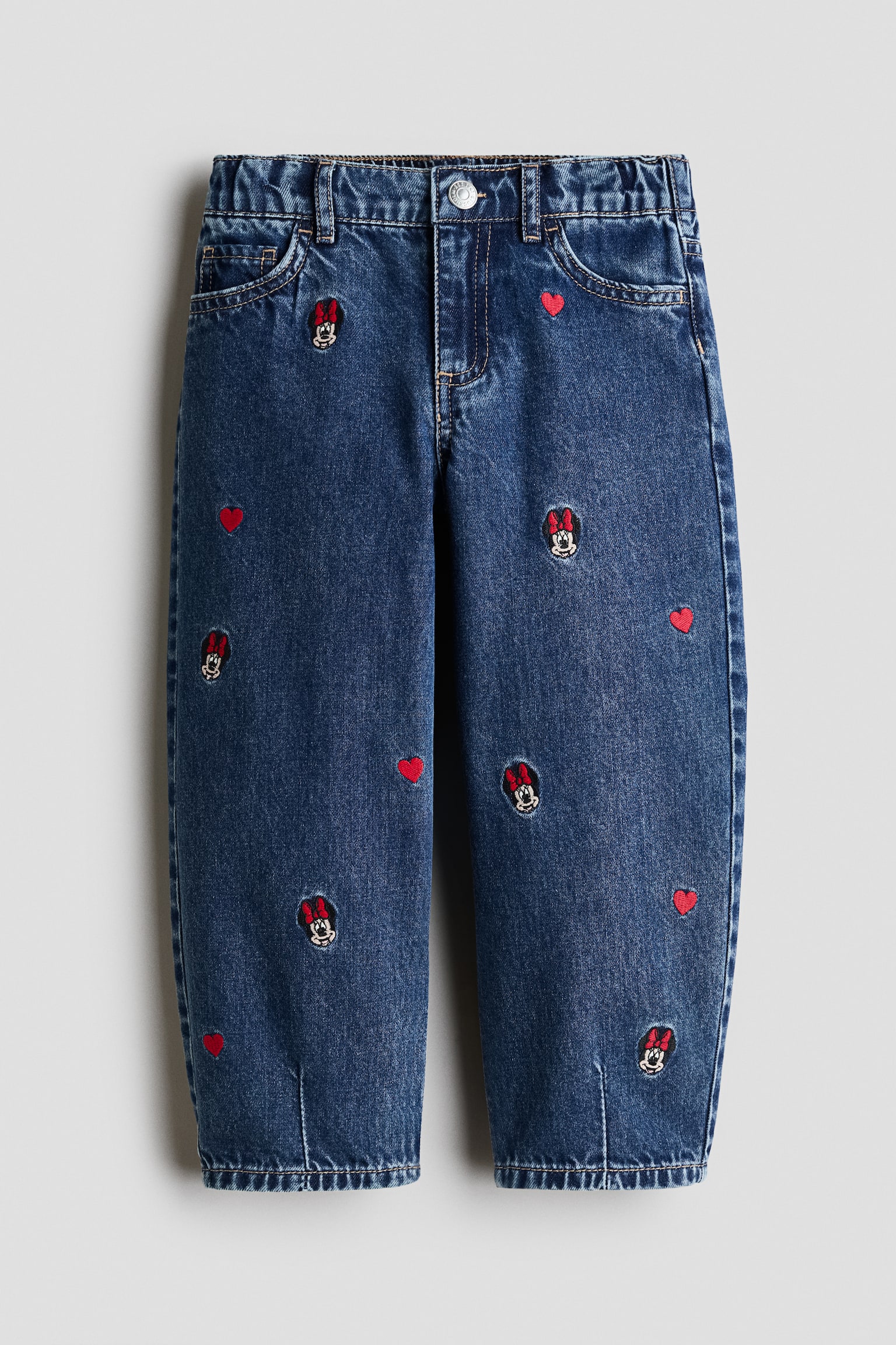 Prikazan je par plavih pamuÄnih denim farmerki sa prednje strane, sa opuÅ¡tenim barrel-leg krojem i vertikalnim uÅ¡ivcima na porubima. Farmerke su ukraÅ¡ene rasutim vezenim glavama Minnie Mouse i malim crvenim srcima na prednjoj strani, a imaju i dizajn sa pet dÅ¾epova, elastiÄni pojas i kopÄanje na pritisak.