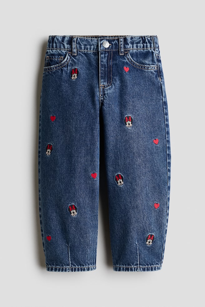 Een paar blauwe katoenen denim jeans wordt van voren getoond, met een losse barrel-leg pasvorm en verticale naden aan de zoom. De jeans zijn voorzien van verspreide geborduurde Minnie Mouse-hoofden en kleine rode hartjes aan de voorkant, en hebben een ontwerp met vijf zakken, een elastische tailleband en een drukknoopsluiting. - 1