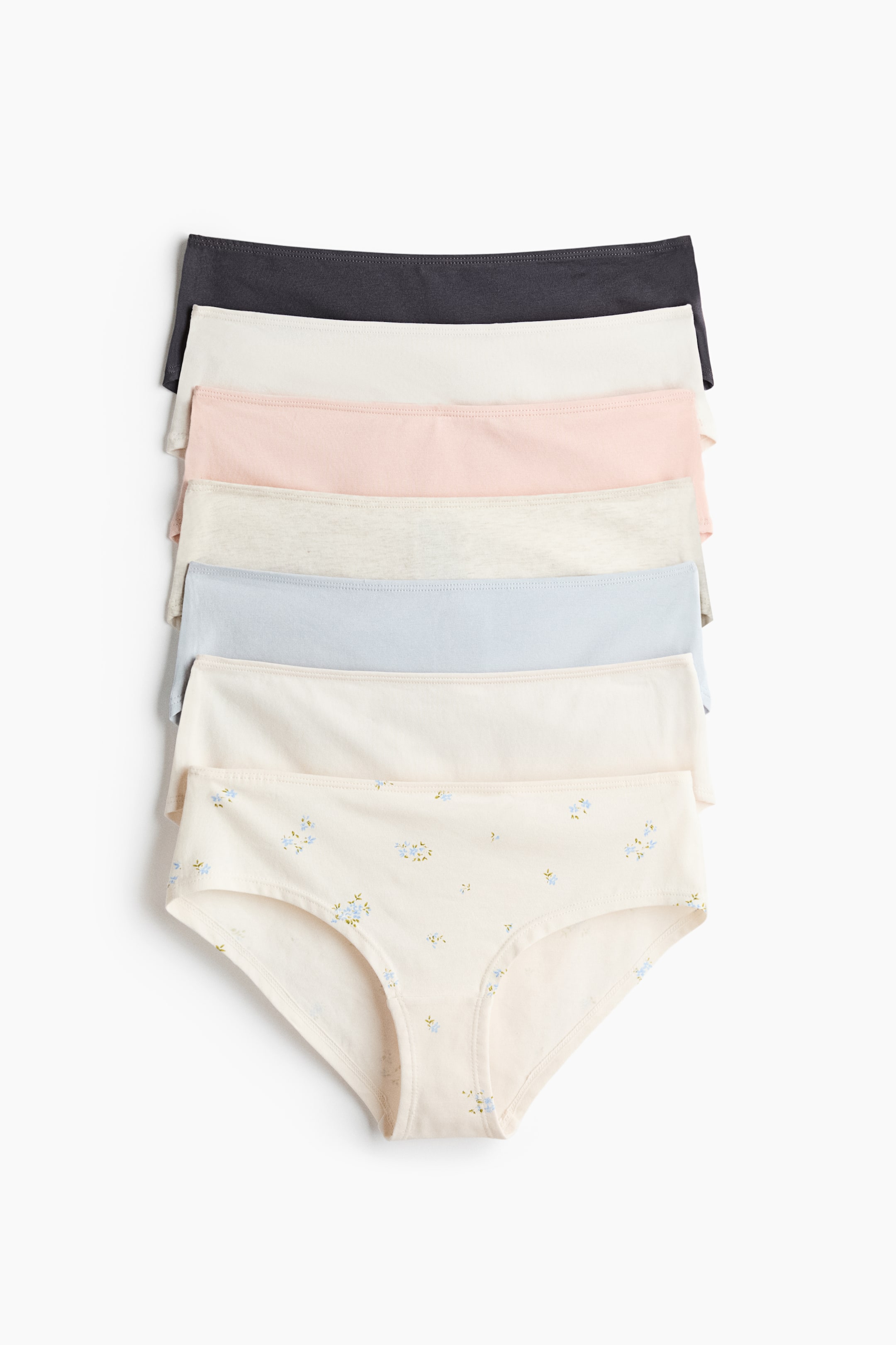Se større billede: En stak af jersey-trusser viser syv par med et design i mellemtalje. De øverste seks trusser er i ensfarvede nuancer, herunder mørkegrå, creme, lyserød, lysegrå og lyseblå, mens den nederste cremefarvede trusse har et lille blåt blomsterprint på den synlige front.